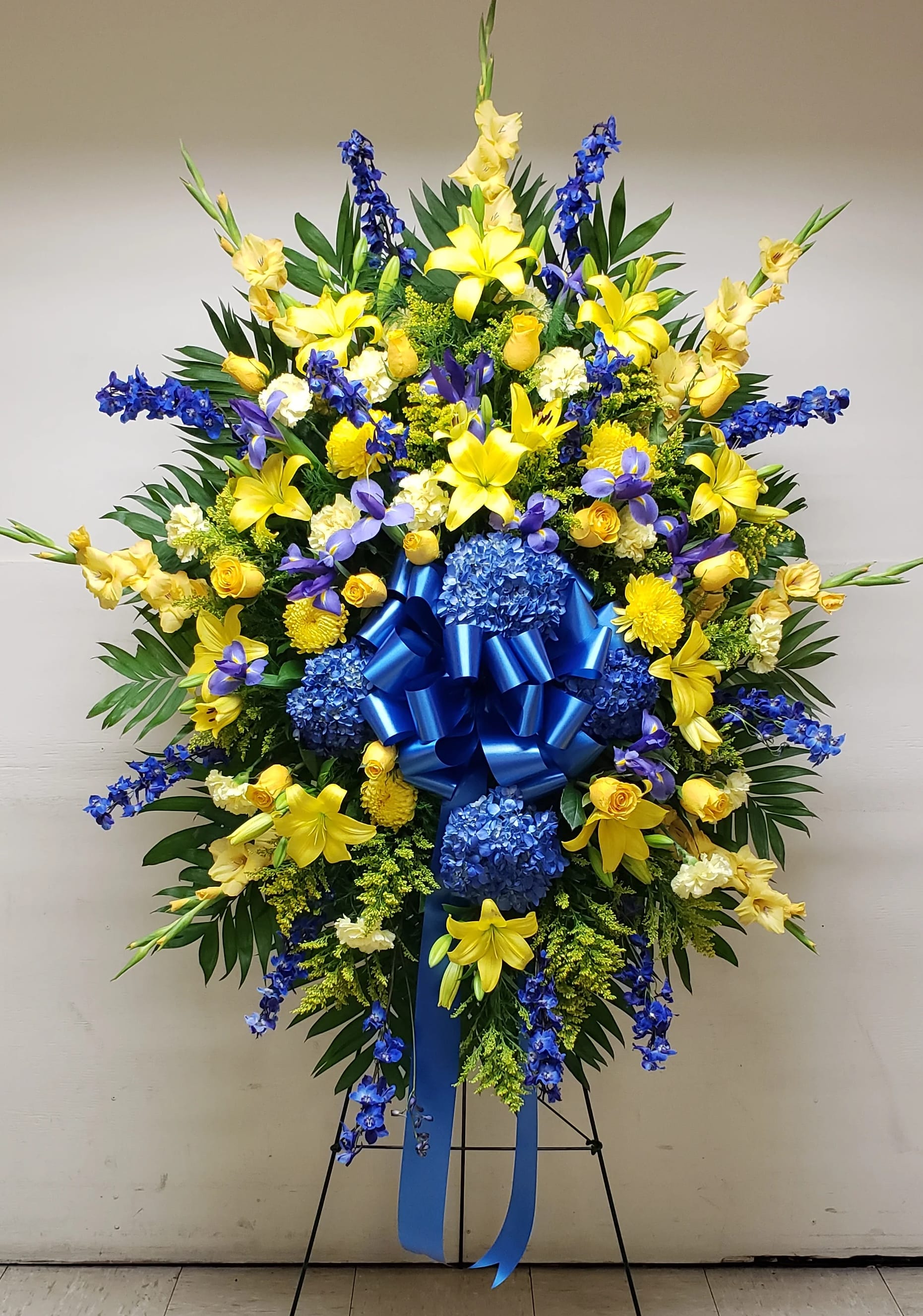 Large standing floral spray with yellow lilies, blue flowers, and a blue ribbon