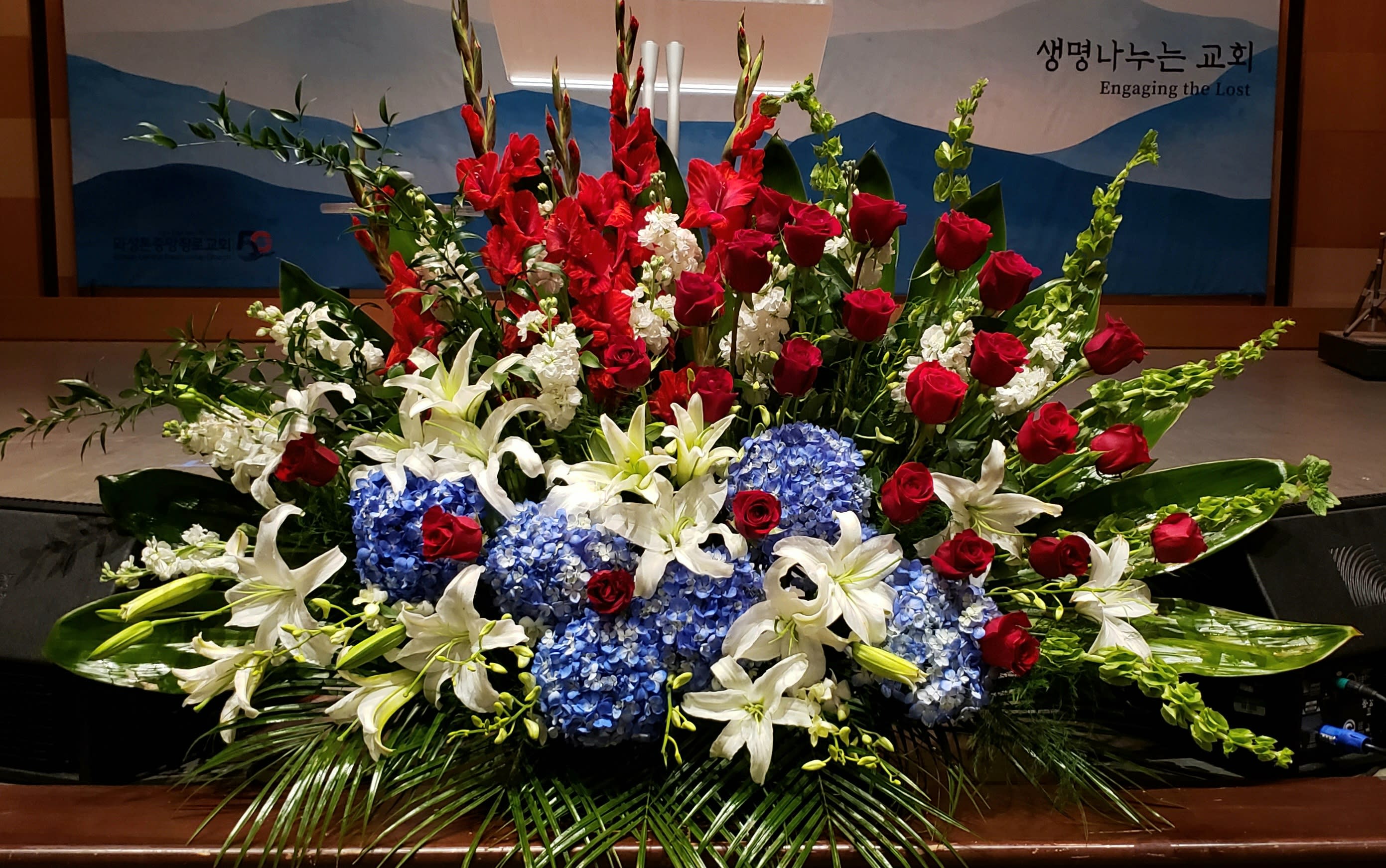 Large red, white, and blue altar arrangement with roses, lilies, hydrangeas, and tall gladiolus.
