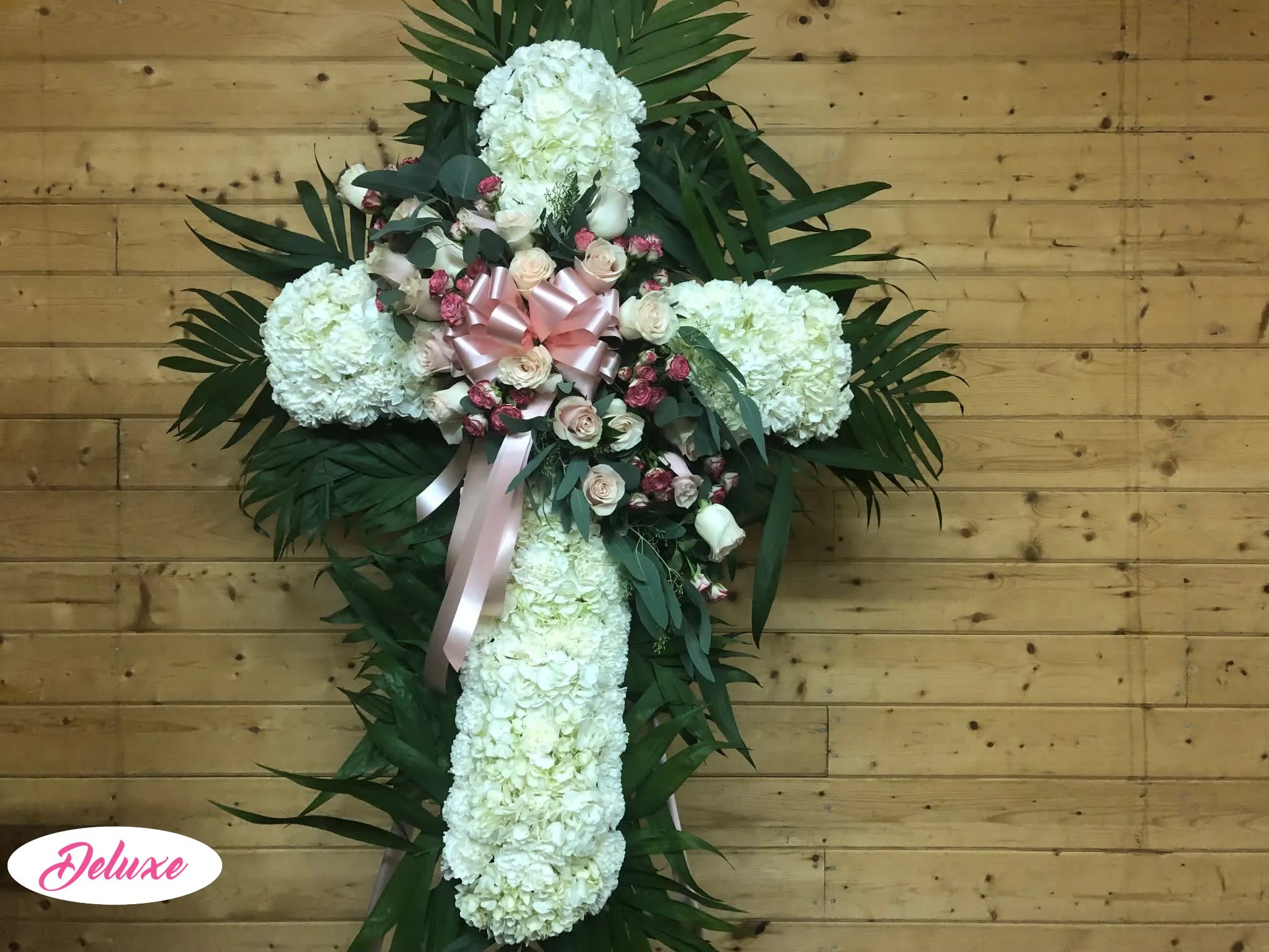 White floral cross arrangement with pink roses and ribbon on a wooden wall