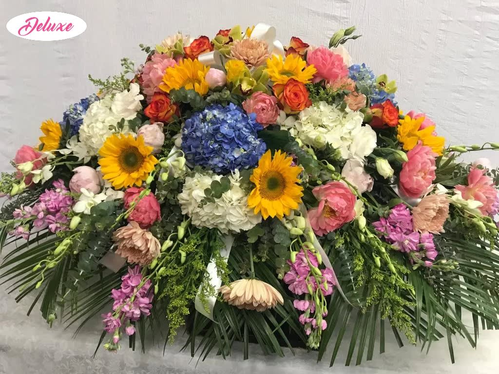 A Garden Paradise Casket Cover  - Garden Paradise is a breathtaking casket cover designed to celebrate a life filled with love, joy, and beautiful memories. Overflowing with vibrant color and natural elegance, this stunning floral tribute features lush blue hydrangea, cheerful sunflowers, soft pink peonies, graceful white orchids, delicate pink stock, and radiant orange roses, thoughtfully arranged to create a garden-inspired expression of love and remembrance. The rich blend of colorful blooms symbolizes hope, admiration, gratitude, and a life beautifully lived, while the elegant white orchids add a touch of peace and timeless grace. Handcrafted by our compassionate florists, Garden Paradise offers a meaningful way to honor a cherished loved one, celebrating their unique spirit while bringing comfort to family and friends during a time of loss.