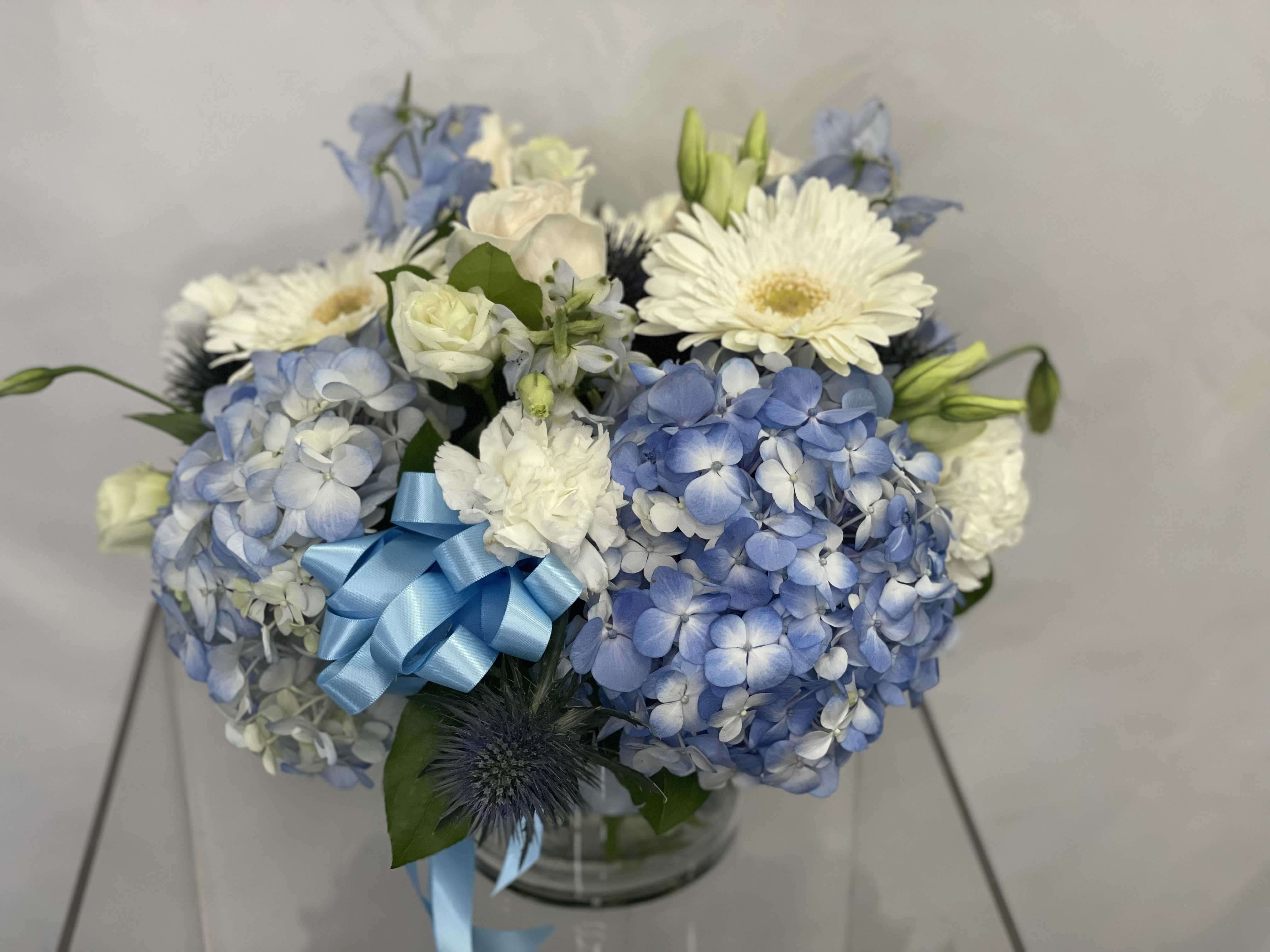 Blue and white bouquet with hydrangeas, gerbera daisies, and a blue ribbon in a glass vase