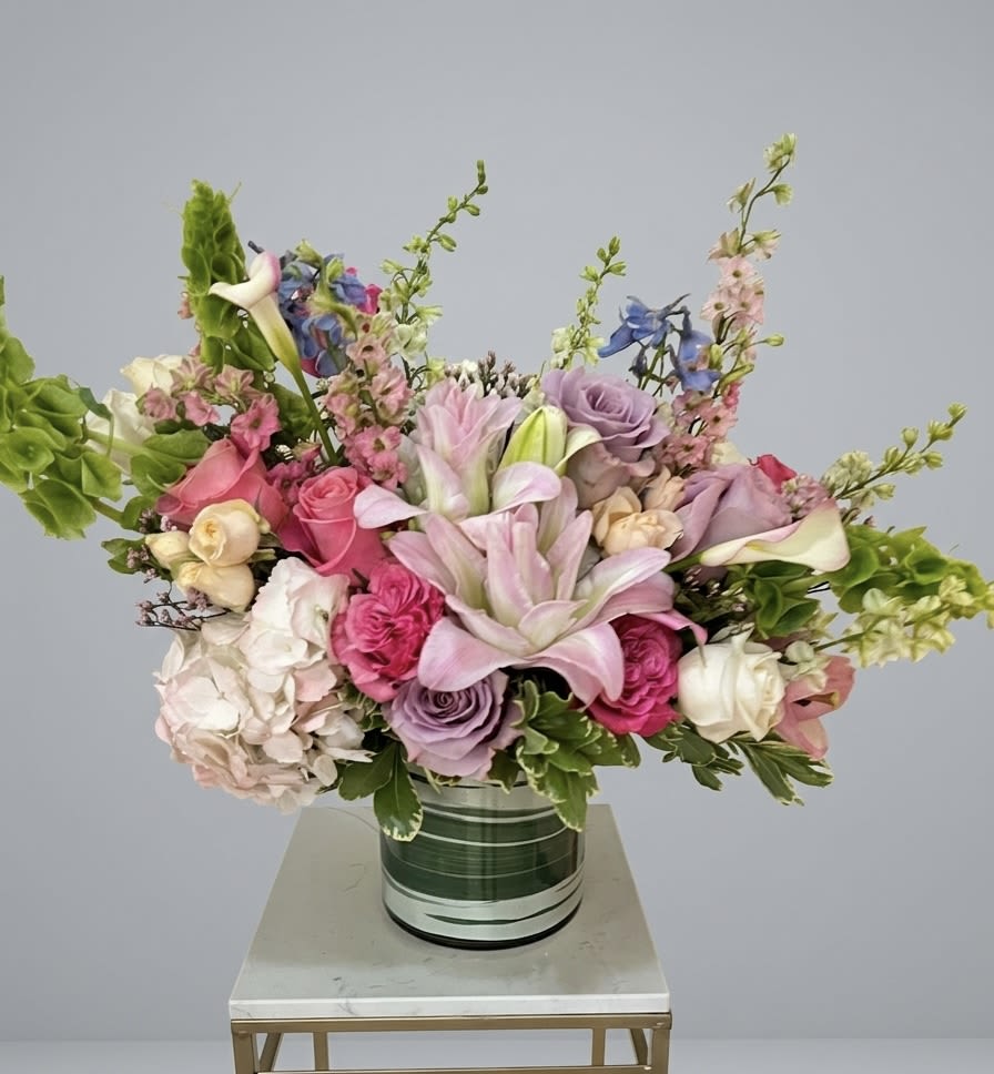 The Grand Opulence Bouquet - The Grand Opulence Bouquet "Indulge in an expression of unparalleled elegance. This striking, masterfully curated arrangement is a masterpiece of texture and refined color. Designed in a sophisticated green and silver cylindrical vase, it sits upon a chic marble and gold accent table, instantly creating a dramatic focal point. Featuring velvety pink and lavender roses, breathtakingly large pink lilies, delicate blue delphinium, and intricate white and cream blooms, this arrangement is a lush symphony of color. Sculptural Bells of Ireland and cascading green foliage add height and movement, capturing a timeless sense of luxury. This is not simply a bouquet; it is an statement of style and sophistication, perfect for elevating any curated interior."