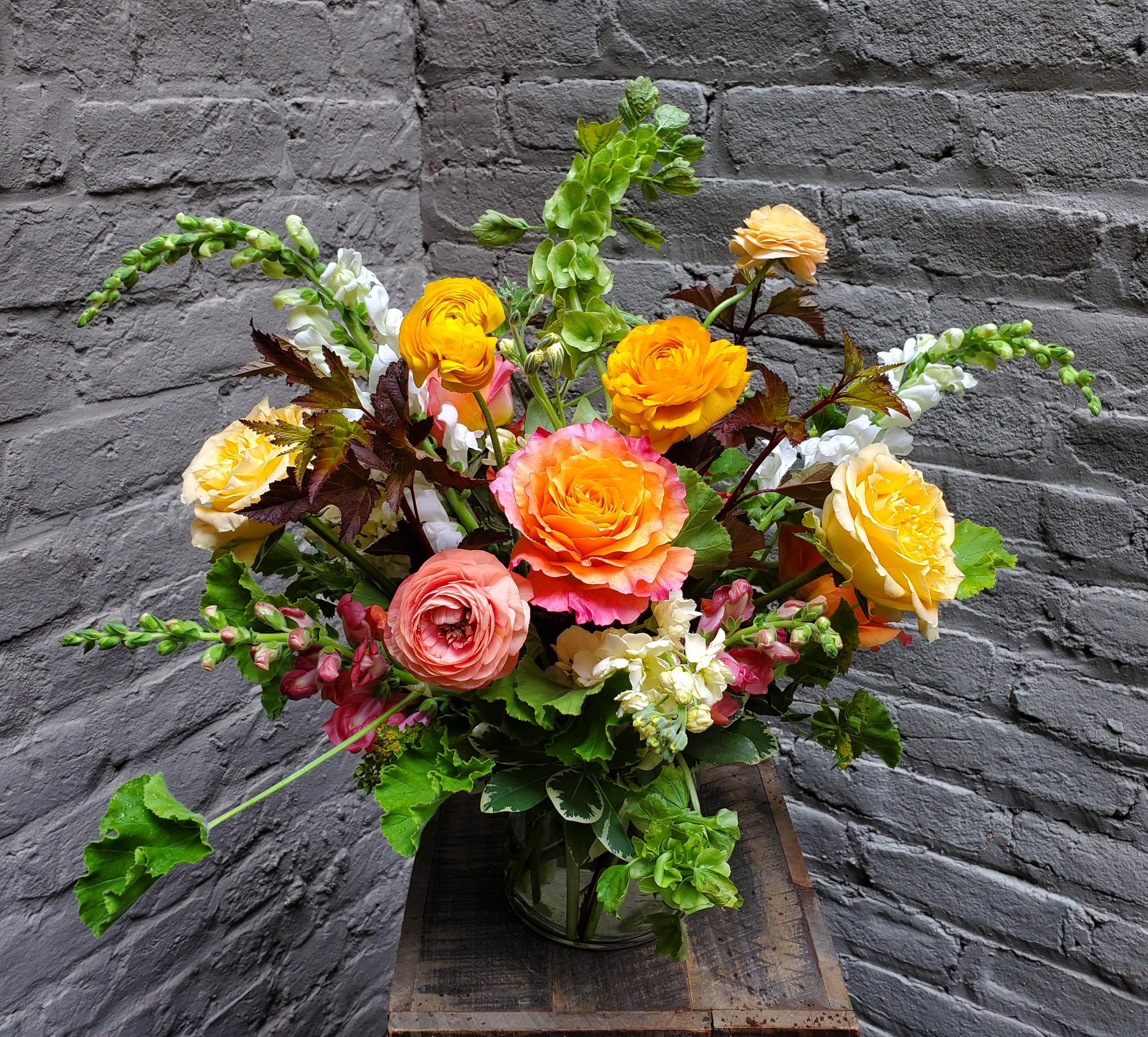 Summer Dreaming - Warm and vibrant, this lush arrangement of roses, ranunculus and snapdragons is the perfect centerpiece for your next summer shindig! Featuring locally-grown foliages and housed in a simple clear glass cylinder vase, it conveys casual, effortless elegance -- the perfect backdrop to a carefree summer evening with the people you love.  Arrangement measures approximately 18" wide by 18" tall.