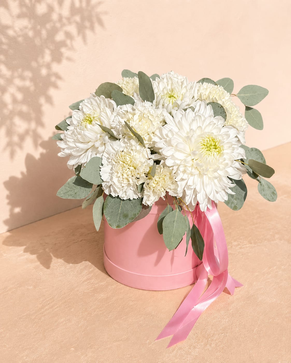 Ariadne's Guiding Star Hat Box Arrangement - Inspired by Ariadne, whose guiding thread became a symbol of hope and direction, Ariadne's Guiding Star celebrates quiet elegance through a refined palette of crisp white blooms and soft silvery greenery.  This sophisticated hat box arrangement showcases premium white chrysanthemums, delicate white carnations, fresh eucalyptus, and seasonal greenery, creating a fresh, airy design with timeless appeal. Presented in Flower Fairy's signature blush hat box and finished with flowing satin ribbons, it offers a beautiful expression of love, gratitude, comfort, and new beginnings.  Perfect for birthdays, sympathy, thank-you gifts, housewarmings, celebrations, or simply brightening someone's day, this elegant arrangement is handcrafted using the freshest premium flowers available.  Flower varieties may vary slightly based on seasonal availability while preserving the arrangement's signature style, color palette, and premium quality.  Features: Premium white chrysanthemums White carnations Fresh eucalyptus Seasonal greenery Signature blush hat box Satin ribbon finish Handcrafted by Flower Fairy Same-day flower delivery in Phoenix
