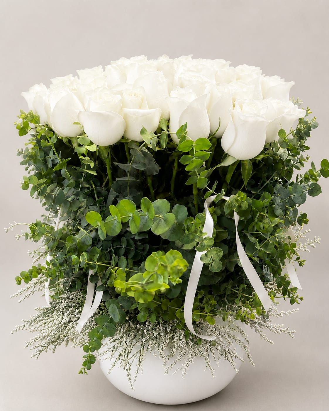 White roses arranged in a round white vase with lush greenery and ribbon accents