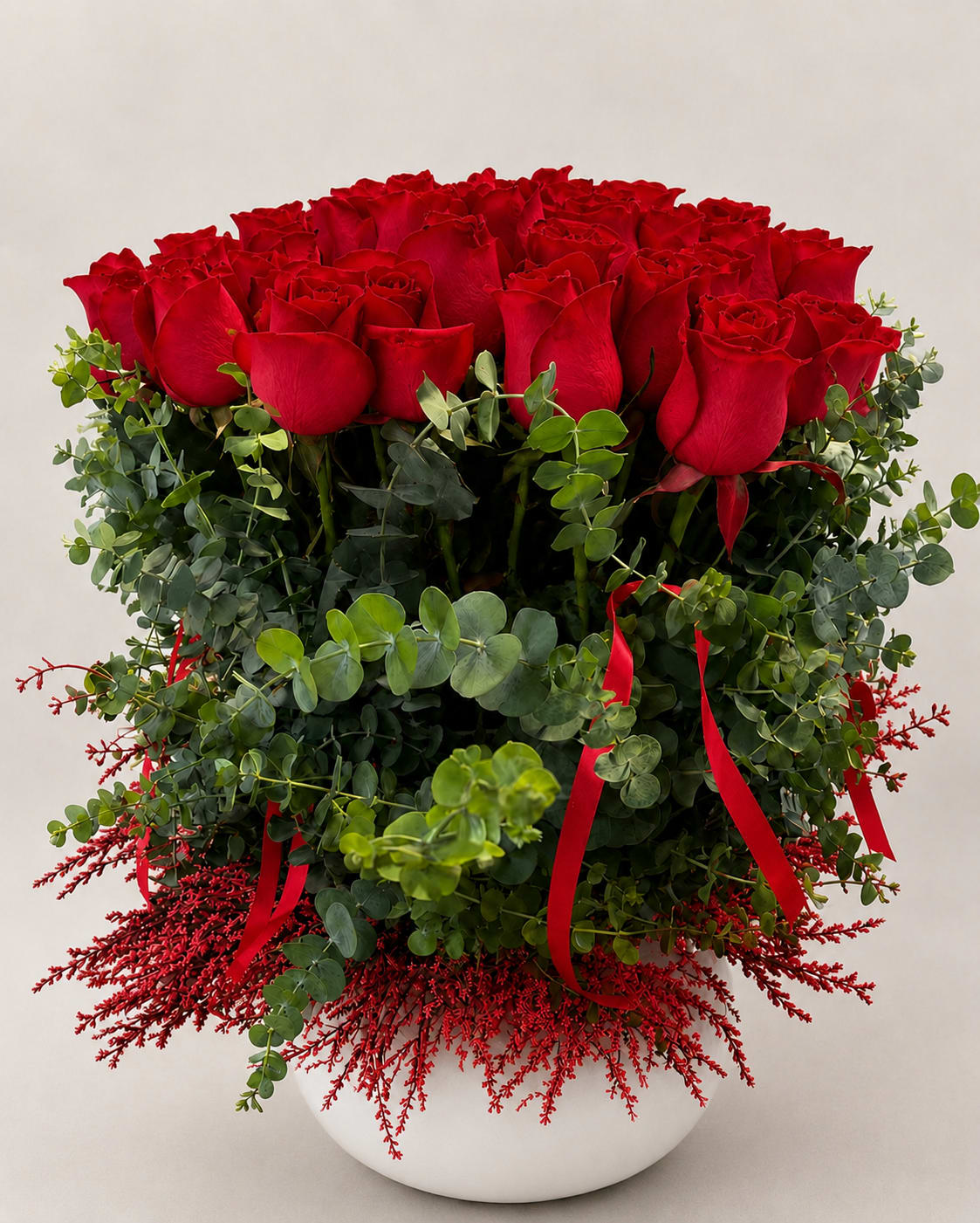 Red roses arranged in a white vase with green foliage and red ribbon accents
