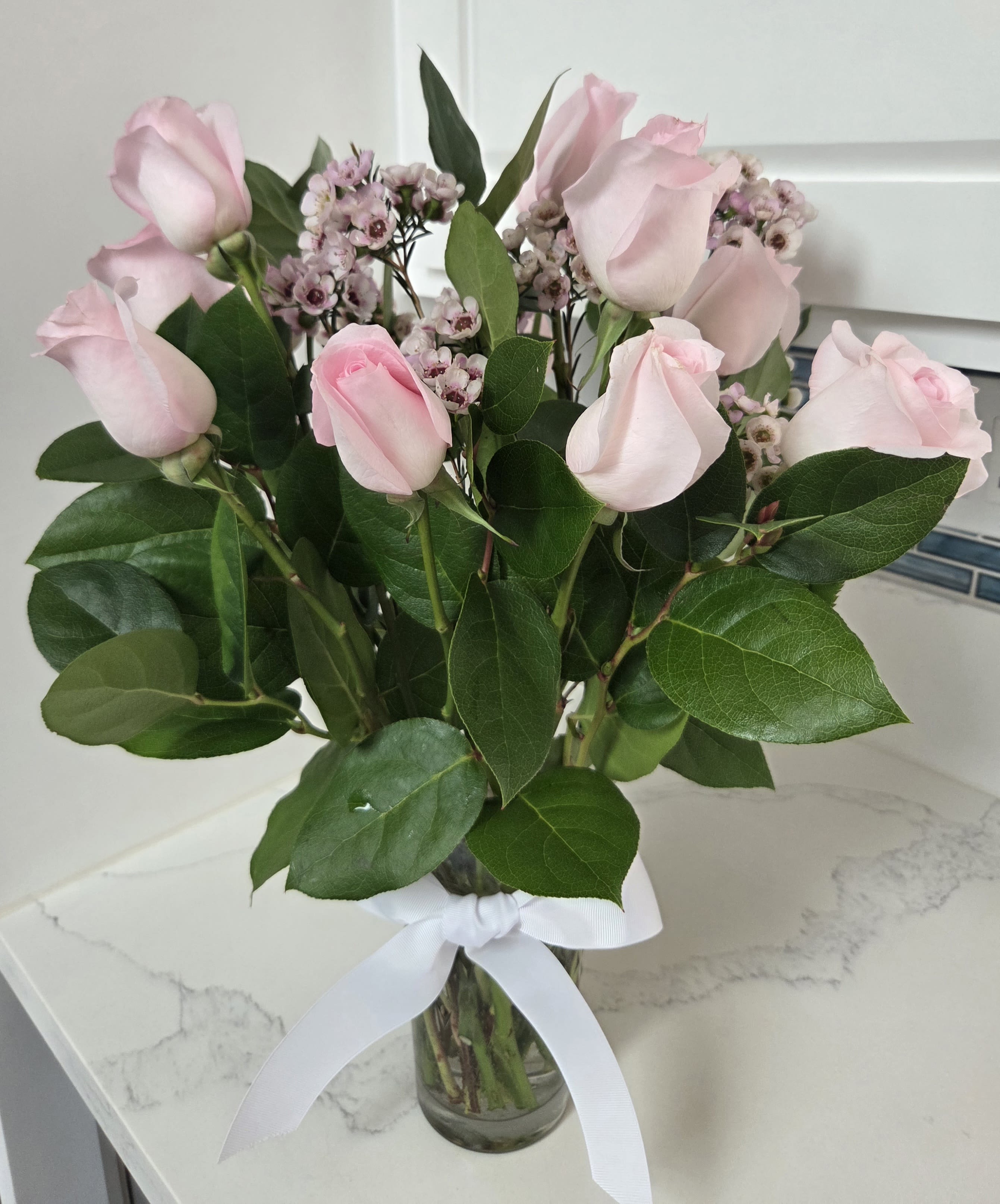 Pink roses and small waxflower blooms in a glass vase with a white ribbon.
