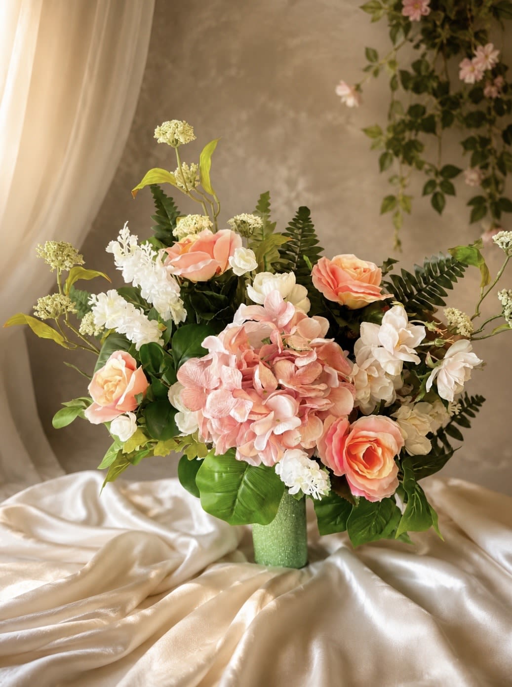 (Silk) Pastel Promise - Soft blush hydrangea crowns this romantic bouquet, framed by large pink roses and delicate white filler blooms-an elegant birthday bouquet or anniversary arrangement that reads both celebratory and tender. The soft pinks and whites create a gentle, comforting impression so she'll feel truly cherished whether you're marking a wedding anniversary, sending sympathy, or brightening an ordinary day with floral warmth.  The display mixes coral-pink and blush roses with a prominent cluster of pink hydrangea, white accent flowers, and glossy green foliage for contrast. The composition is full and layered with a wide, balanced silhouette in one version and a loose, garden-style asymmetry in the other, giving it a natural, freshly-cut look that fits a variety of décor styles.  This arrangement suits recipients who appreciate a classic romantic palette-mothers, partners, or close friends-offering a thoughtful way to say "I'm thinking of you" or "congratulations." Vase included; stems are arranged for immediate display and will benefit from fresh water every few days and a clean vase to keep blooms looking their best.