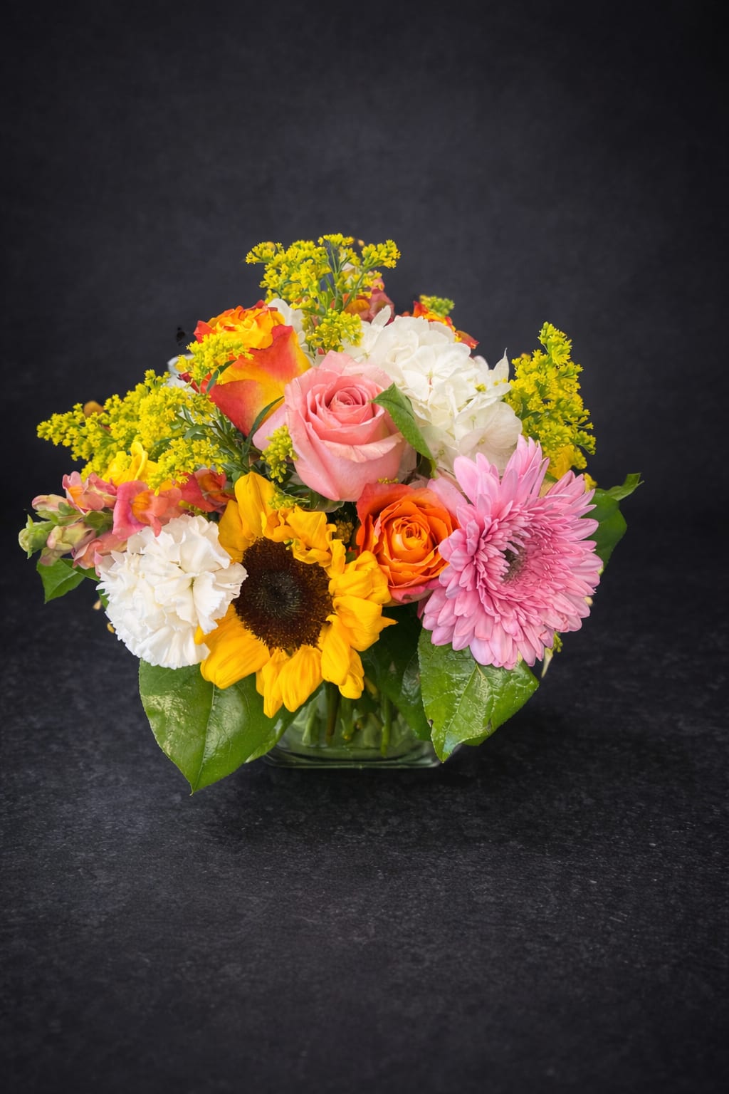 Mixed bouquet with roses, sunflowers, carnations, and pink daisies in a glass vase
