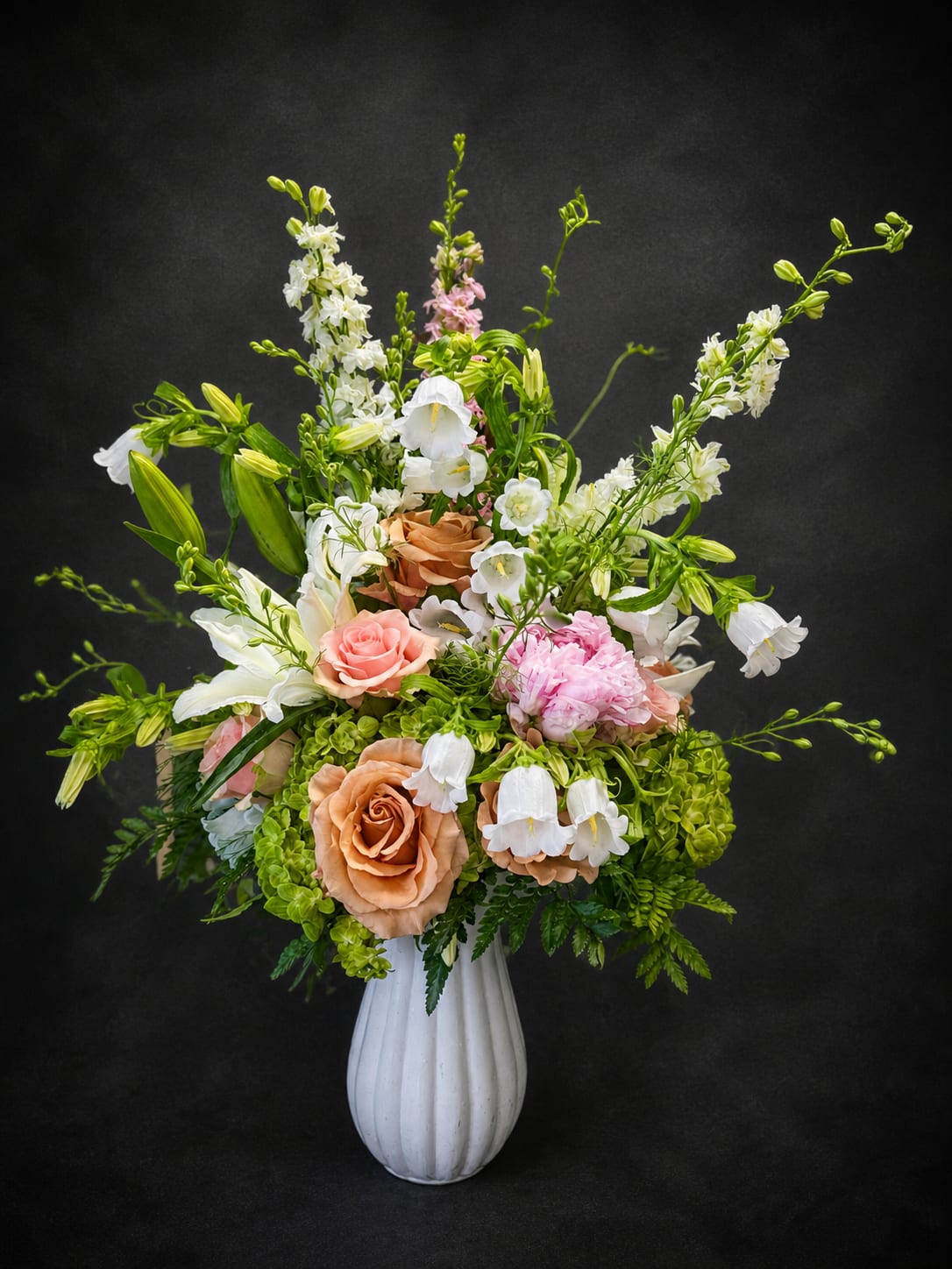 Peaches and Cream - Peach roses and trumpet lilies rise in a tall, garden-inspired arrangement that feels both elegant and effortlessly romantic - perfect as a birthday bouquet, anniversary flowers, or a thoughtful everyday gift. The airy silhouette and soft pastel palette of peach, pink, white, and green create a gentle impression that will make the person you're thinking of feel truly seen and celebrated.  This design pairs long-stemmed snapdragons and lilies with clusters of peach roses and delicate white bellflowers, all set in a ribbed white ceramic vase. The composition is built with taller stems above a dense base of blooms for graceful movement and visual height; the white vase adds a refined, modern touch that complements the soft colors. The mix reads fresh and garden-like, with a light, romantic texture ideal for a mantel, entry table, or dining centerpiece.  Sized to make a statement without overwhelming a room, this arrangement suits celebrations, sympathy, or simply brightening someone's day. Arrives ready to display in the included white ceramic vase - minimal care needed beyond fresh water and a clean vase to keep the blooms looking their best.
