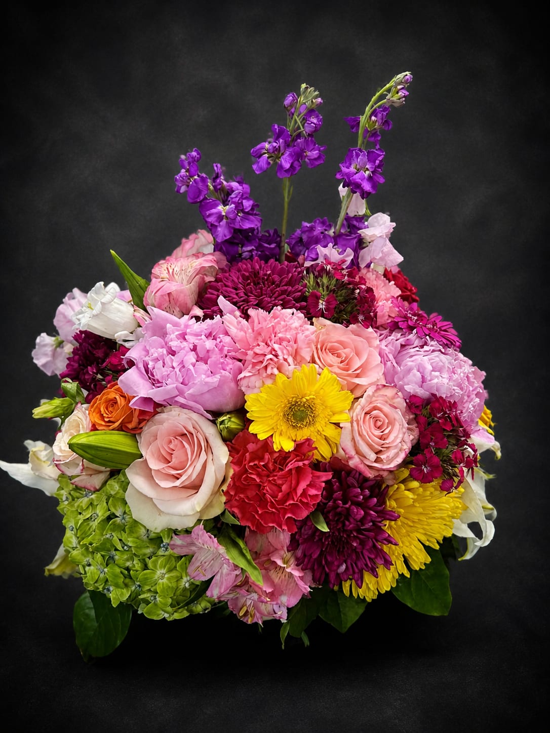 Diva’s Delight - A lively, garden-fresh bouquet that reads like a joyful celebration-Diva's Delight is a mixed flower arrangement perfect as a birthday bouquet, thank-you gift, or cheerful spring surprise. The rounded, densely layered design places fragrant lilies and roses amid ruffled carnations and chrysanthemums, with tall purple stock rising above the cluster to add height and drama so the recipient feels truly celebrated.  Colors pop in pink, purple, yellow, orange, and white for a feminine, upbeat palette; alstroemeria and other filler blooms weave texture and contrast through the center for a full, textured look. The composition is arranged close and compact for immediate visual impact, giving a lush, freshly gathered-from-the-garden impression that brightens any room.  Choose this arrangement when you want someone to feel noticed and adored-whether marking a birthday, sending thanks, or lifting spirits on an ordinary day. Care is simple: keep blooms in fresh water, trim stems every few days, and display away from direct heat to extend their life.