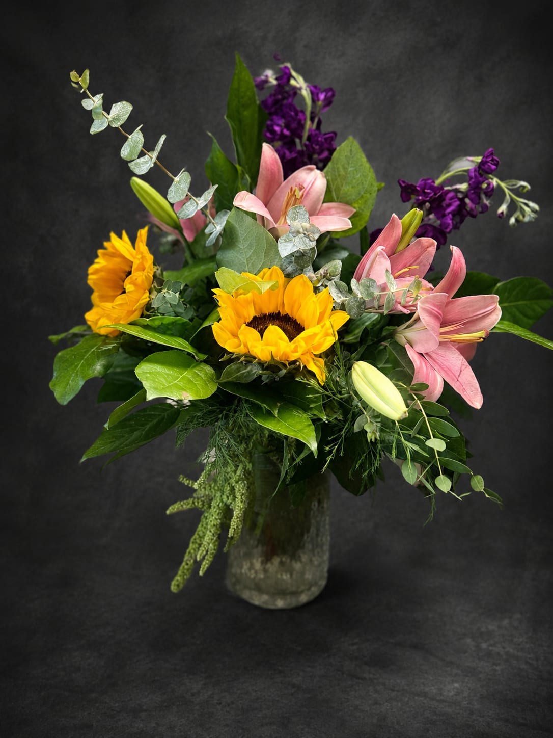 Sunshine Wishes - Bright yellow sunflowers and soft pink lilies create an immediate, sunny welcome in this mixed-color flower arrangement-perfect as a birthday bouquet, a thank-you gift, or a spring celebration centerpiece. The asymmetrical, full design pairs bold warm tones with deep purple accents so the person you're sending it to feels noticed and joyful the moment it arrives.  Arranged in a clear glass vase, the bouquet blends large sunflower heads and layered lily blooms with airy eucalyptus-like stems and glossy greenery for height, texture, and movement. The color story-vibrant yellow, blush pink, and cool purple-adds cheerful contrast and a touch of drama without being fussy.  Choose this arrangement when you want someone to feel genuinely celebrated or uplifted; it's the kind of everyday bouquet that brightens a kitchen table or office desk. Vase included; no special care needed beyond fresh water and a cool spot away from direct heat to keep blooms looking their best.