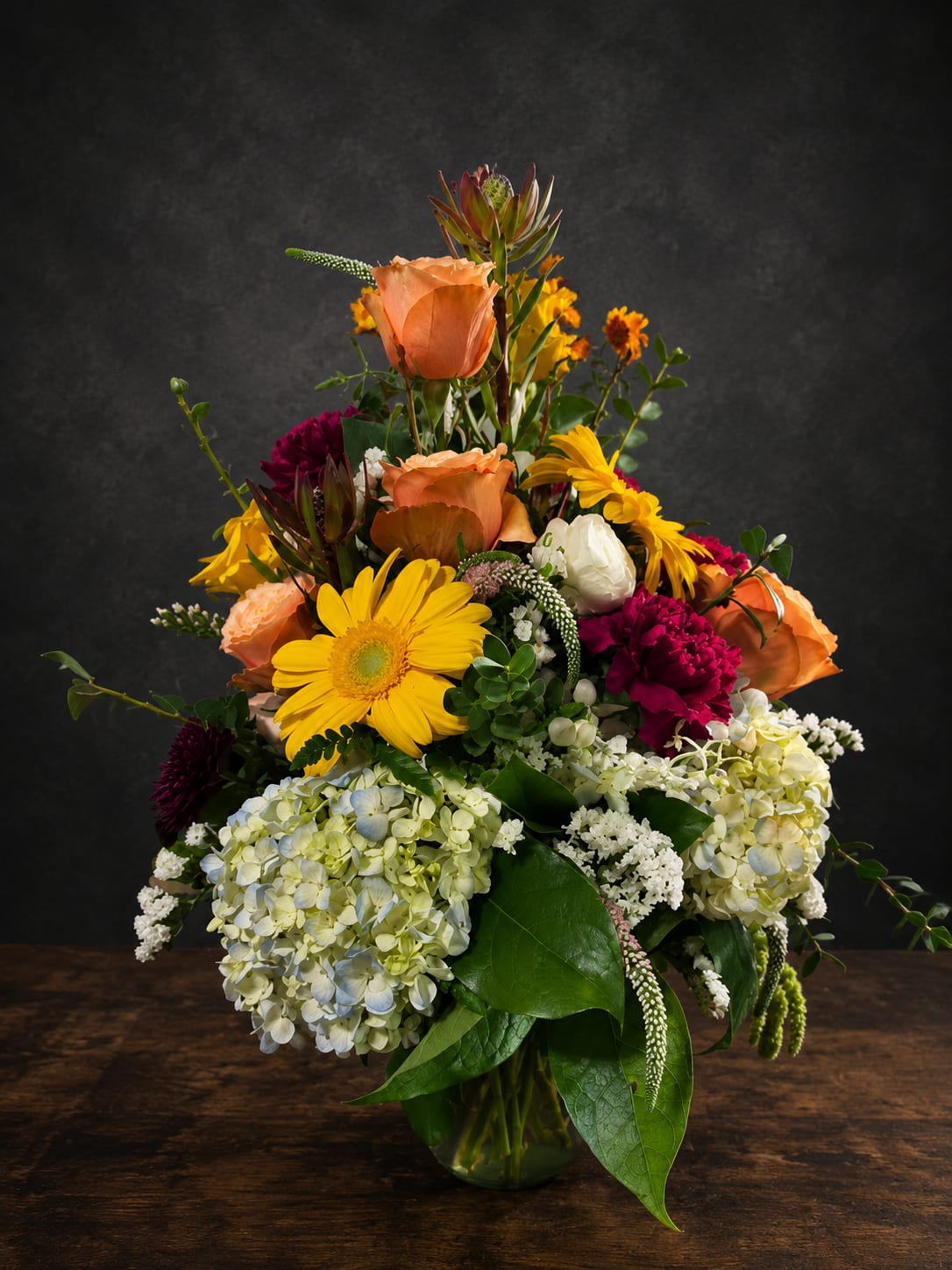 Southern Charm - Bright yellow gerbera daisies and soft peach roses create an immediate burst of cheer in this mixed color flower arrangement - the perfect birthday bouquet or thank-you gift that looks freshly gathered from a sunlit garden. Tall accent stems and a loose, layered shape give the arrangement height and movement so the person you're thinking of will feel truly celebrated the moment they see it.  This full, garden-style arrangement pairs large clusters of pale white hydrangeas with deep magenta carnations, daisy-like yellow blooms, and textured filler flowers for lively contrast and a lush, abundant silhouette. Arranged in a clear glass vase, the combination of yellow, pink, white, peach, and orange tones and glossy foliage reads bright and festive from every angle.  Ideal for birthdays, spring occasions, congratulations, or simply to say "I'm thinking of you," Southern Charm arrives ready to display in the included glass vase. Its varied heights and sturdy stems make it easy to enjoy - keep water fresh and trim stems occasionally to prolong the cheerful look.