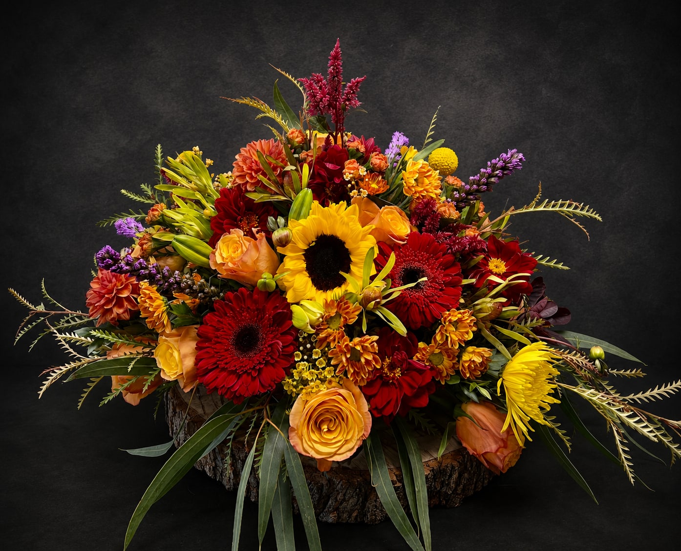 You Light Up My Life - A bold sunflower anchors this vibrant fall-inspired flower arrangement-perfect as an anniversary bouquet or a radiant birthday centerpiece. This "You Light Up My Life" mixed floral arrangement reads like a celebration in color: sunflowers and red gerbera daisies deliver sunny warmth, while orange roses, deep dahlias and chrysanthemums add rich, autumnal depth with purple and lavender accents that catch the eye. It's the kind of arrangement that makes the person you're thinking of feel noticed and truly celebrated.  Arranged densely in a rustic bark container, the composition is low and abundant with layered blooms and long, arching foliage that create movement and a full, rounded silhouette. Textured accents and clustered smaller flowers fill the vase for a lush, tactile look; colors include bright yellow, deep red, warm orange, purple and green for a festive, energetic palette. Sized as a large, statement piece, it works beautifully as a dining table centerpiece or a striking gift display.  Ideal for many occasions-graduation, housewarming, get well wishes, thank yous, "just because" moments, or to say "I'm sorry"-this arrangement also carries the warmth suited to spring and summer celebrations as well as sympathy displays when richer tones are preferred. Care is simple: keep the container watered and in a cool spot away from direct midday sun to extend bloom life; available in a large, full design to brighten any room or meaningful moment.