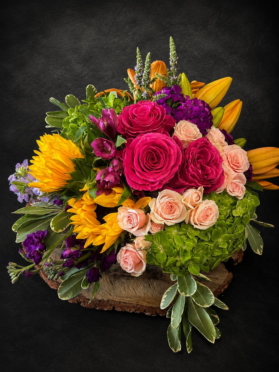 Love You So Much - Hot-pink roses and bright yellow sunflowers take center stage in this garden-style flower arrangement for anniversary bouquets, birthdays, or just because surprises. The vibrant mix-magenta roses, orange lilies, peach spray roses and clusters of purple accents-creates an energetic, celebratory iimpression that says "you matter" to the person you're sending it to, whether you're congratulating, saying thank you, or cheering someone who's under the weather.  Built dense and low on a rustic wood slab base, the composition also includes green hydrangea and broad variegated foliage for lush contrast and layered texture. The rounded, slightly asymmetrical shape reads abundant and fresh; sizes and proportions are designed to sit out on a table or mantel as a focal piece. Care is simple: keep water fresh, trim stems every few days, and place away from direct heat to enjoy the vivid colors longer.  This arrangement suits a wide range of moments-housewarming, get well, sympathy or funeral remembrances handled with bright warmth, Mother's Day, or a thoughtful "I'm sorry" or "thinking of you." Its bold color story and sturdy wood base make it a confident, cheerful gift that makes the recipient feel genuinely celebrated.