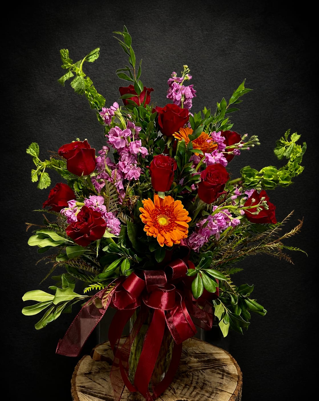 The Rose Affair - Deep, velvety red roses pair with bright orange gerbera daisies and soft pink stock to create a bold, romantic flower arrangement perfect for an anniversary, birthday, or "thinking of you" surprise. The tall, garden-style composition reads dramatic and celebratory-the person you're sending this to will feel noticed and adored whether it's for Valentine's Day, Mother's Day, a graduation, or simply because you miss them.  This full, upright design is layered with abundant mixed greenery and long, arching accents for movement and texture, finished with a burgundy ribbon tied around a clear glass vase for a polished presentation. Expect a lush mix of roses, gerbera daisies, stock (and complementary blooms), arranged to showcase high-contrast reds, oranges, pinks, and fresh greens in a show-stopping silhouette.  A versatile gift for occasions from get well and housewarming to retirement, sympathy, or an "I'm sorry" gesture, this arrangement arrives ready to display in its vase. Choose sizing at checkout for the right impact; simple care-fresh water and a cool spot-will keep the blooms looking radiant through spring and summer and beyond.