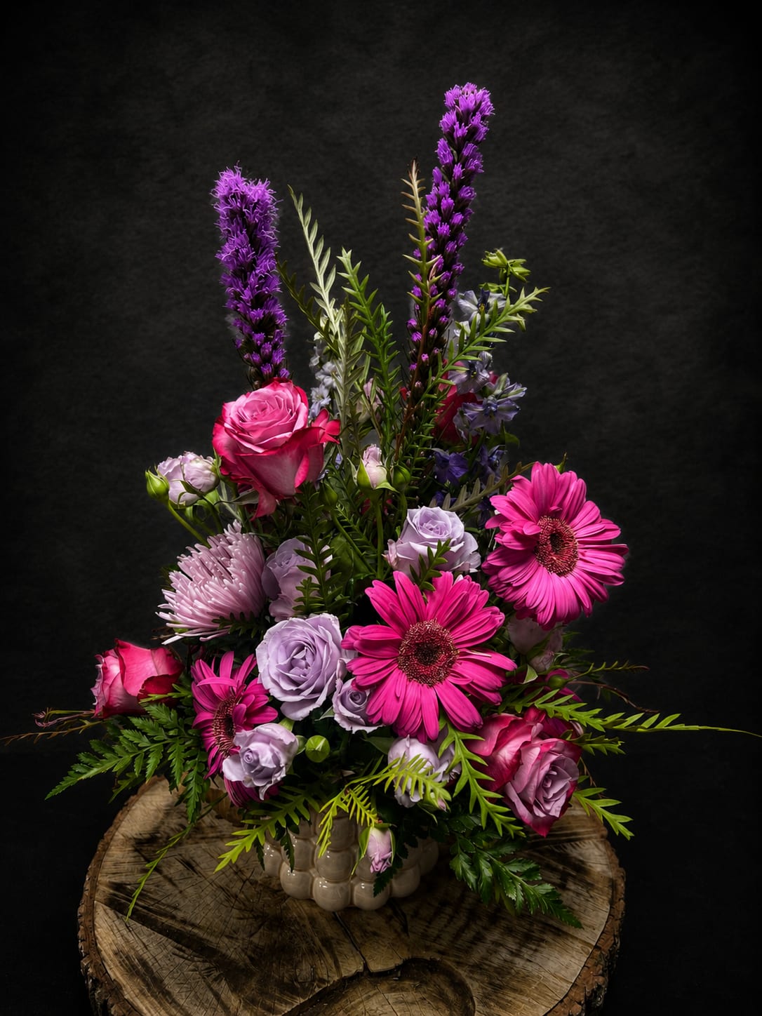 Lavender Mist - Bright pink gerbera daisies and roses meet soft lavender roses and pale lisianthus in a lively anniversary bouquet that reads romantic and modern at once. Tall purple spiked blooms rise above the dense lower cluster to create a dramatic, upright silhouette-perfect as an anniversary bouquet or a memorable birthday, graduation, or "thinking of you" gift that will make the recipient feel truly celebrated.  This full arrangement layers saturated pinks, soft lilacs, white accents and fresh green foliage for contrast and texture. Presented in a rounded ceramic vase, the design stands about upright and fills a tabletop or entryway with color and presence; the combination of gerbera, roses, lisianthus and snapdragon-like spikes gives a mix of round blooms and vertical movement that reads elegant yet cheerful.  Ideal for occasions from birthdays, Mother's Day, Valentine's Day and graduations to get-well wishes, retirements, thank-you gestures, sympathy, "I'm sorry" or simply "just because," Lavender Mist is a versatile gift that comforts and delights. For best results, keep the vase topped up with fresh water and away from direct heat-perfect for sending to someone you want to make feel seen and cared for.