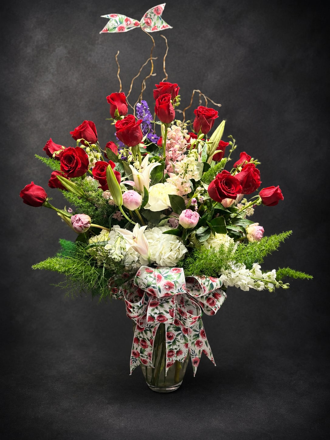 Cherry Blossom  - Bright red roses rising above white lilies and soft pink blooms give this tall vase arrangement an immediately romantic, celebratory presence-perfect as an anniversary bouquet, Valentine's Day gift, or a special birthday surprise. The layered, airy silhouette and purple accent flowers add depth and contrast so the person you're sending it to will feel truly seen and celebrated.  Built on a dense base of white hydrangea-like clusters and fresh greenery, the design features numerous red roses, white lilies, pale pink flowers, and touches of purple for visual pop. It arrives in a clear glass vase wrapped with a floral-print ribbon and finished with matching decorative butterfly and floral picks for a polished, gift-like presentation. The composition is tall and full, with blooms rising above a broad, layered profile that makes a striking focal display.  This arrangement works beautifully for romantic moments, milestone birthdays, Mother's Day, or to offer sympathy with grace-its formal yet whimsical accents suit a range of sentiments. Care is simple: keep vase water fresh and trim stems every few days to extend bloom life; available in the tall vase style as pictured for maximum impact.