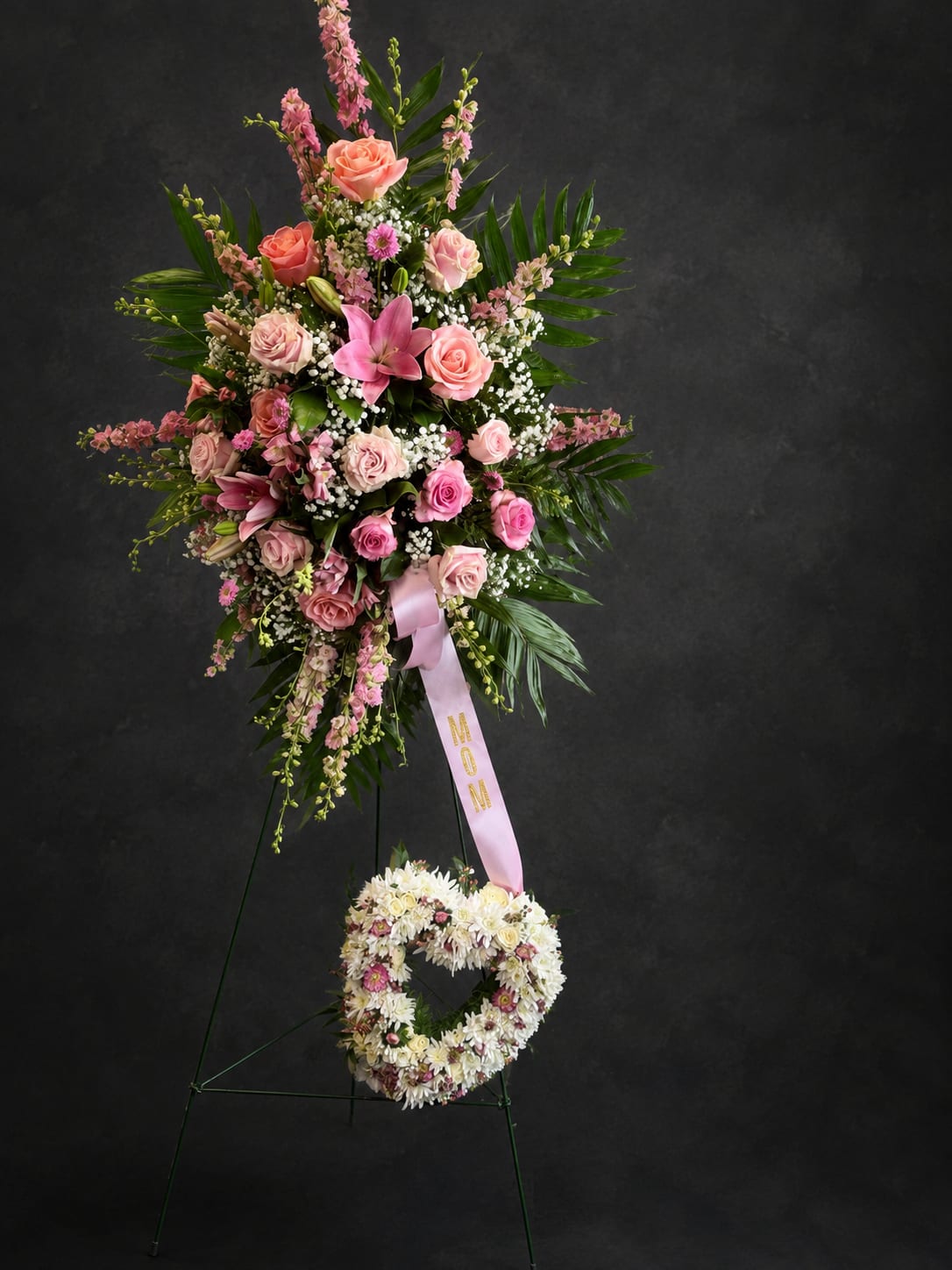 With All My Heart - A tall, heart‑felt standing spray that reads as a warm and personal tribute - perfect as a sympathy arrangement or a memorial for Mother's Day. The design pairs a generous upper spray of pink roses and a prominent pink lily with soft white filler blooms and lush dark green, palm‑style foliage, while a matching heart‑shaped wreath sits below on the metal easel. A pale pink ribbon printed with "MOM" gives the composition an intimate, commemorative touch that comforts and honors the person you're remembering.  The arrangement blends roses, lilies, carnations, chrysanthemums and assorted pink and white blooms for a layered, formal silhouette that looks graceful from every angle. Finished on a sturdy metal floral easel, the piece measures as a traditional standing tribute with a complementary floral wreath beneath; textures range from the silky rose petals and lily blooms to the denser mums and clustered filler flowers, creating depth and a lush visual impact. This is the kind of tribute that lets the recipient feel truly seen and remembered - please note the included pink ribbon and easel are integral to the presentation when ordering.