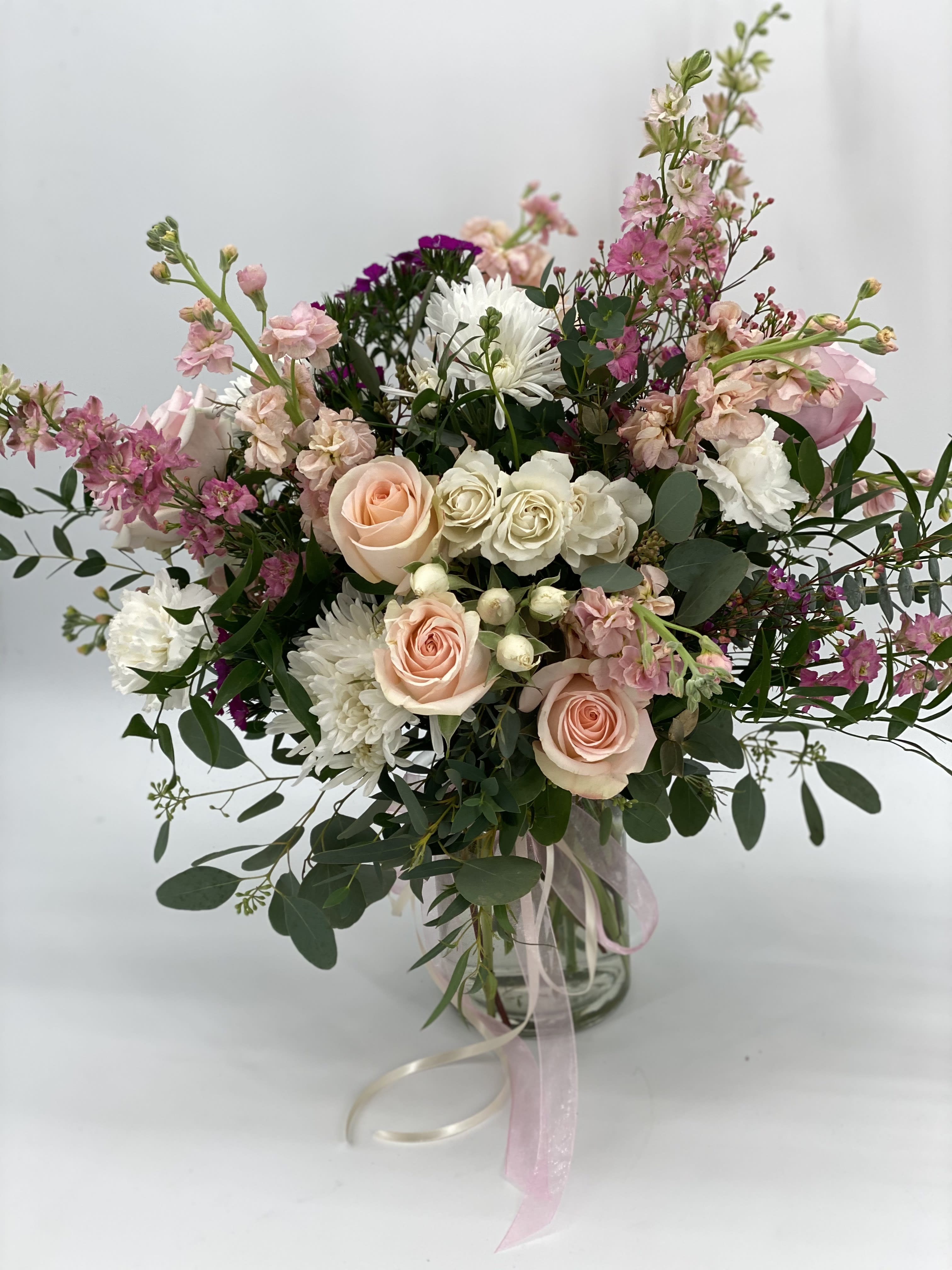 Tall arrangement of peach roses, white mums, and pink blooms in a glass vase with trailing ribbons
