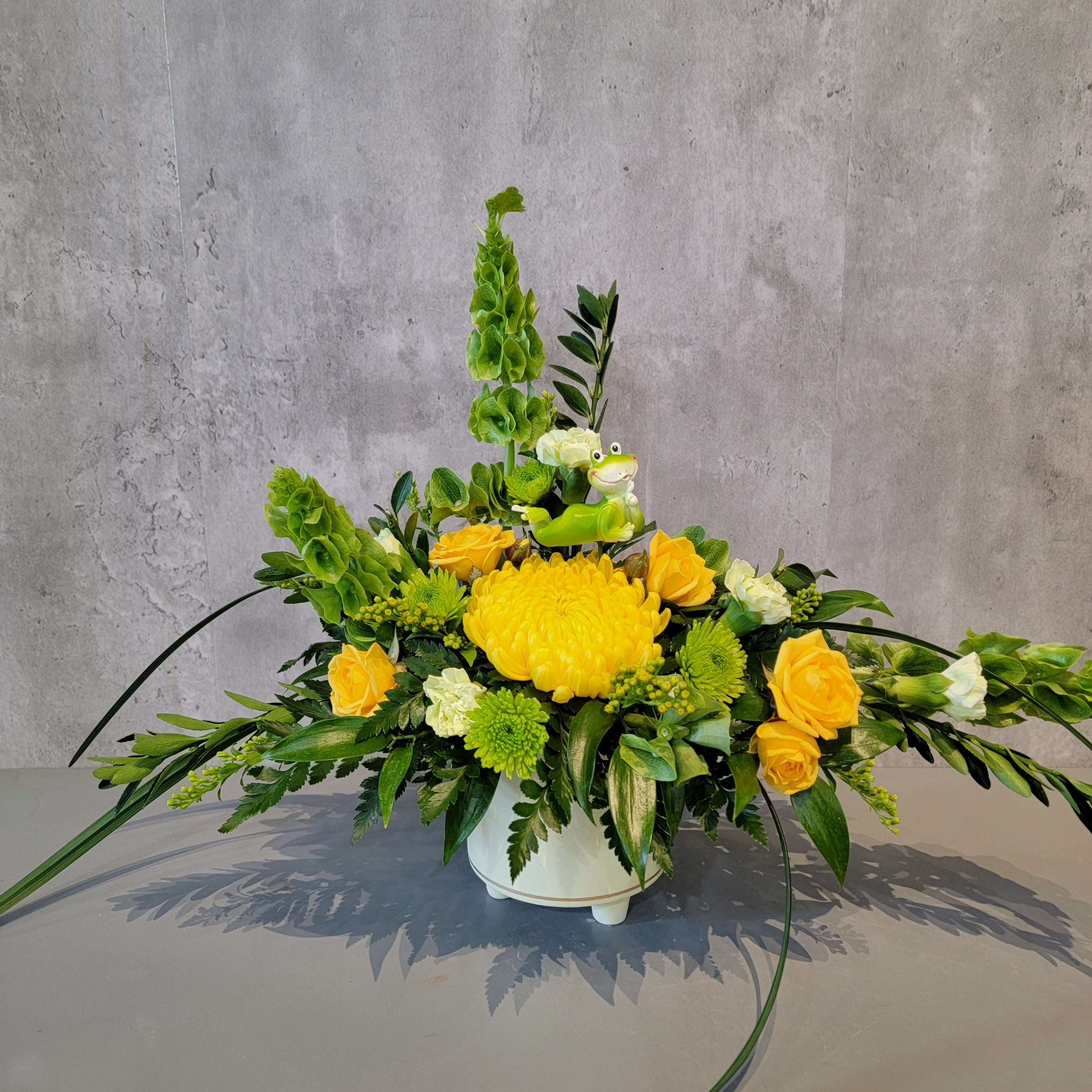 Yellow and green floral arrangement in a white container with a small frog figurine