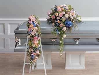 Pastel flower casket spray with matching cascading arrangement beside a silver casket