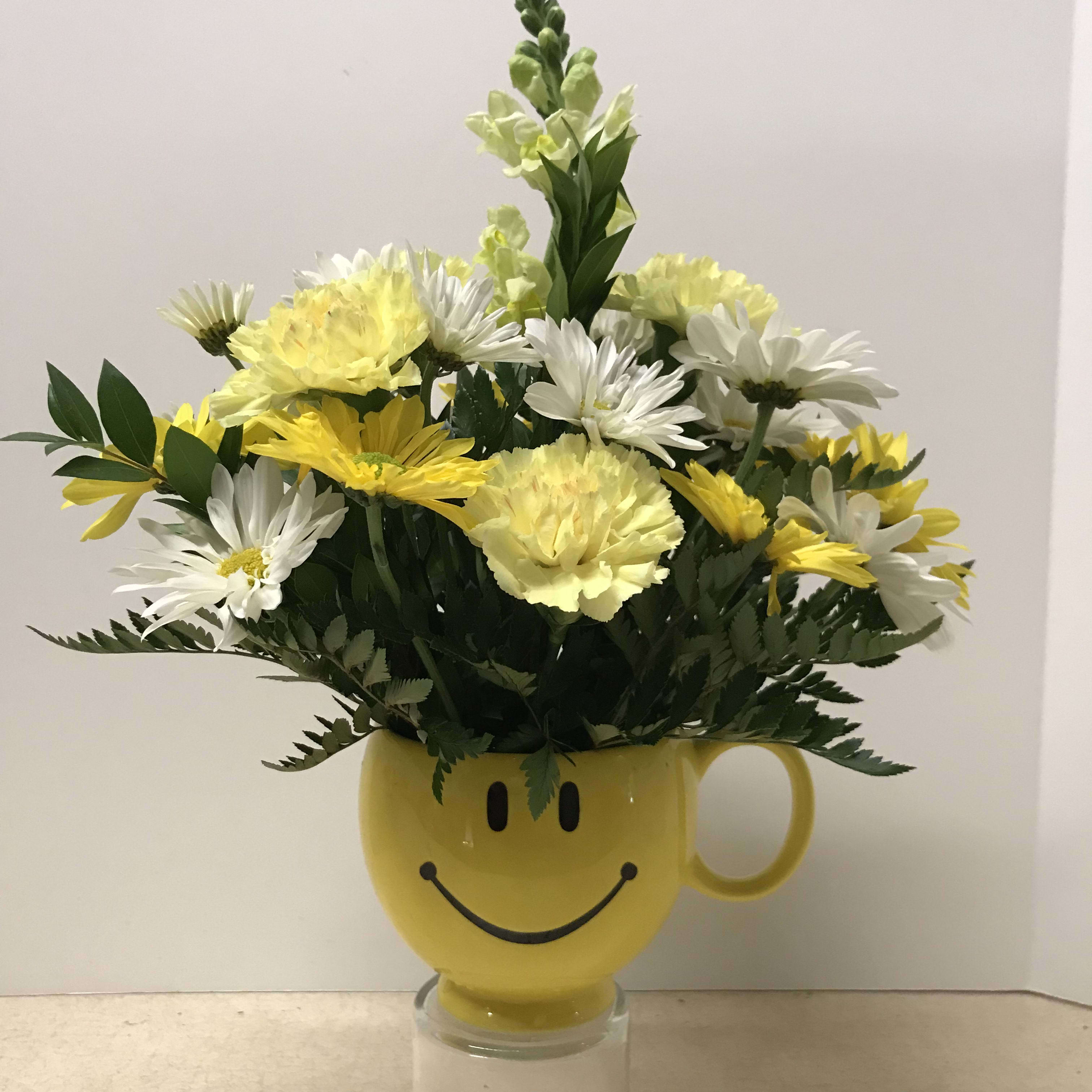 "Sunshine and Smiles" - A cheerful arrangement in a smiley container!  Filled with yellow and white daisies, carnations, snapdragons, and assorted greens.  A perfect way to bring a smile to anyone!  Why not gift one today to your friend, family member, or loved one?  