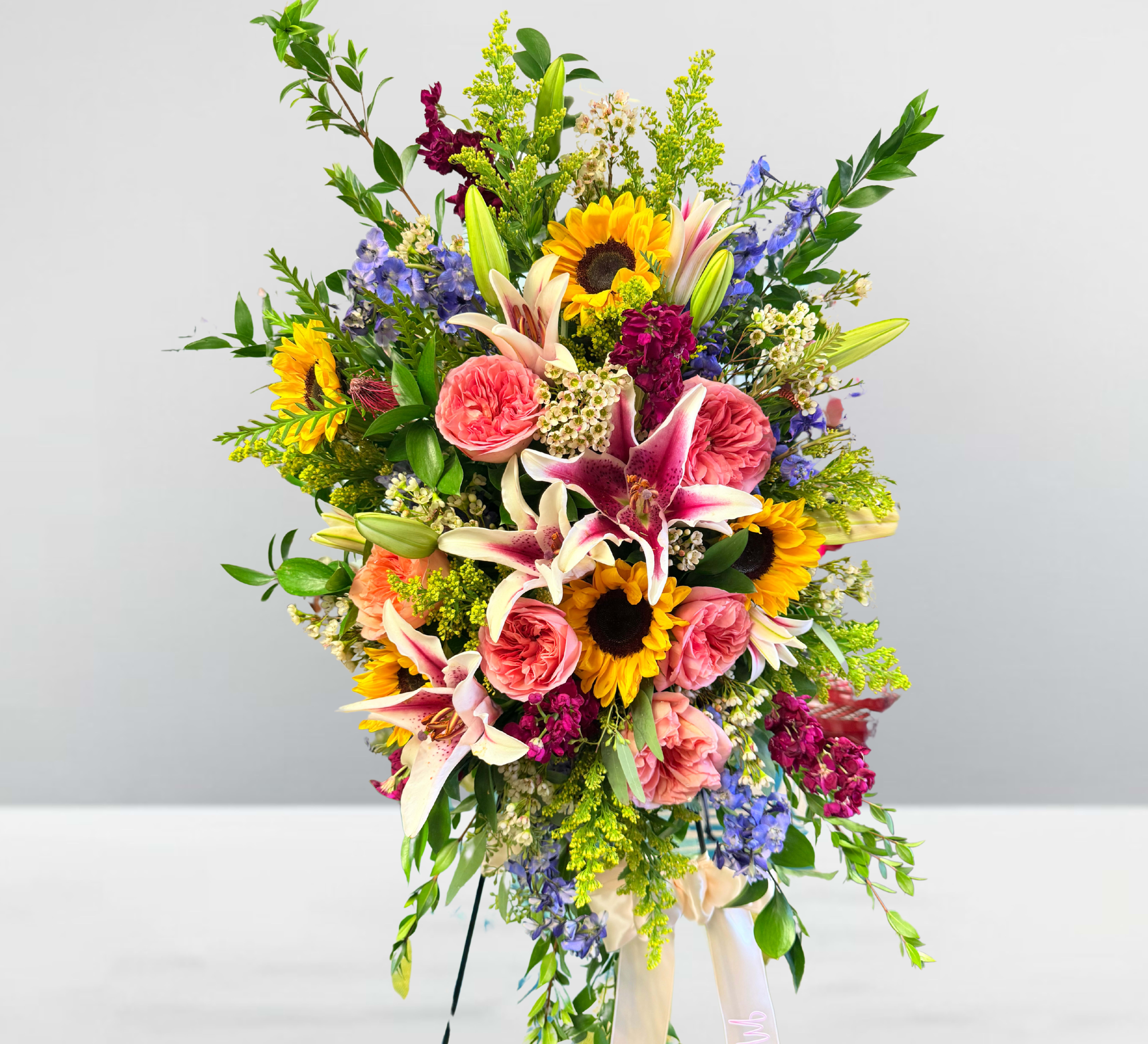 Sincerely loved standing spray  - Sincerely Loved Standing Spray  Express your deepest sympathy and heartfelt love with the Sincerely Loved Standing Spray, a stunning tribute filled with vibrant beauty and warmth. This exquisite arrangement showcases graceful Stargazer lilies, elegant pink Expression roses, and radiant sunflowers, symbolizing love, admiration, and remembrance. Delphinium and stock add delicate texture and height, creating a lush and breathtaking display.  With its rich colors and carefully chosen blooms, this standing spray serves as a beautiful farewell, honoring a life sincerely loved and deeply cherished. Perfect for funerals, memorial services, or any moment of remembrance, it offers comfort and beauty during times of loss.  Let this floral tribute speak from the heart and celebrate a life filled with love and joy.