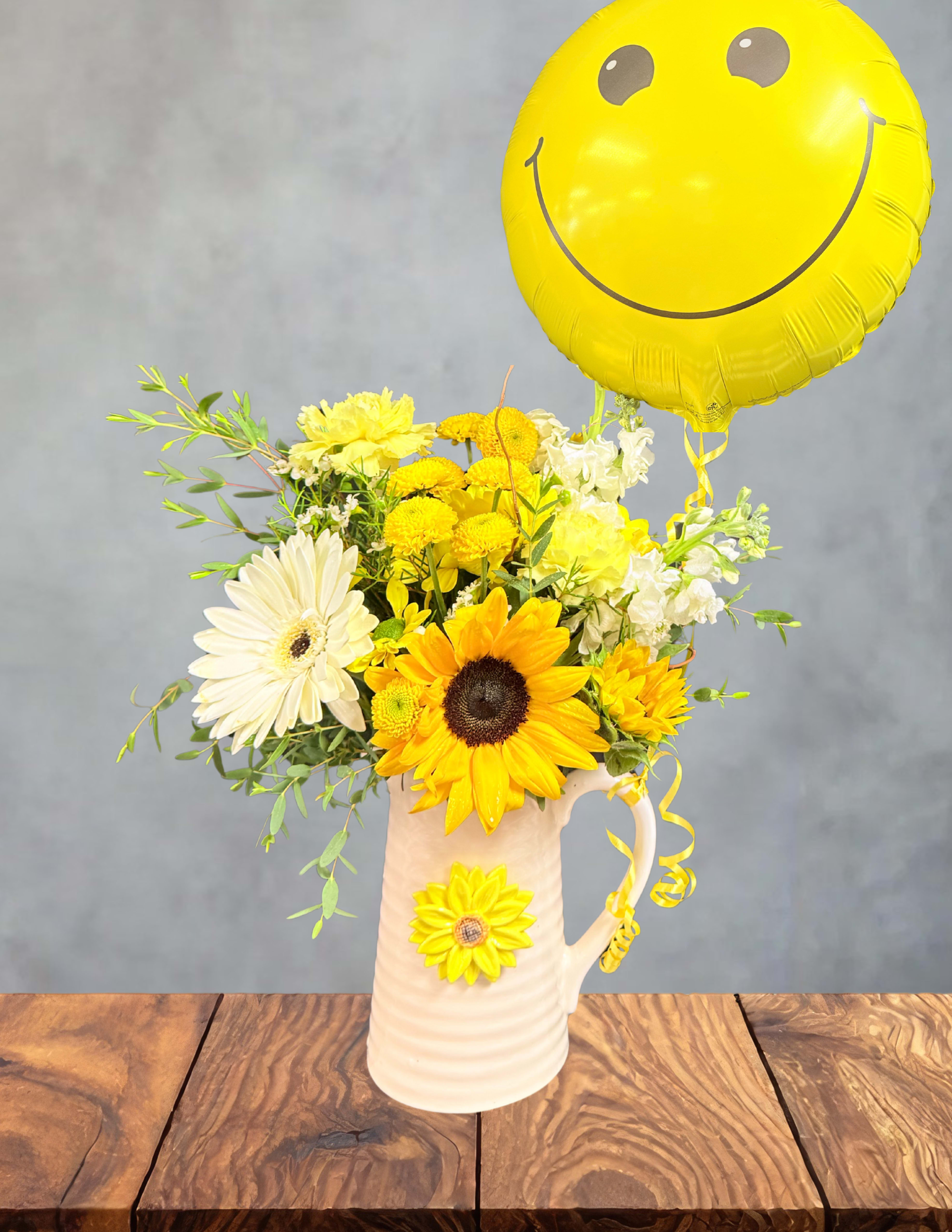 Yellow and white flower arrangement in a pitcher with a yellow smiley balloon