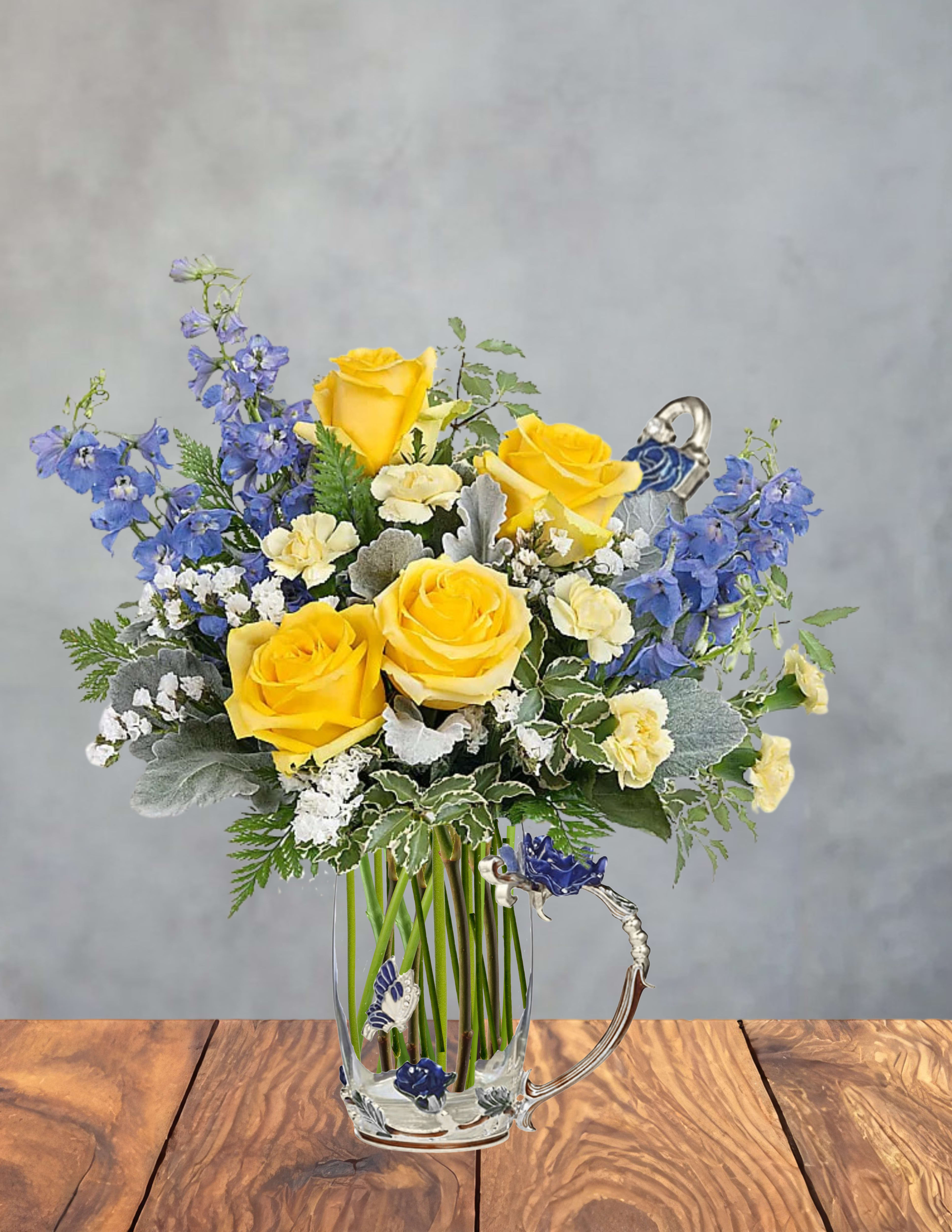 Bright Hugs Bouquet - Bright yellow roses form the sunny heart of this flower arrangement, making it an ideal birthday bouquet or cheerful pick-me-up for someone who deserves a bright moment. Blue delphinium spikes and cream carnations fan out from the center, while silvery foliage and delicate white filler flowers add garden-style texture and soft contrast-perfect for saying "thank you," "thinking of you," or celebrating an everyday success.  The flowers arrive arranged in a clear glass mug with a decorative handle, turning the arrangement into a keepsake gift that's both pretty and practical. Included decorative cup accessories-glossy enamel-like charms in blue, purple and gold shaped like a rose and a butterfly with small rhinestone accents-give the presentation a playful, feminine touch alongside the metal spoon. The palette of yellow, blue, white, green and purple creates a fresh, uplifting look that will brighten a desk, kitchen counter, or bedside table.  This is the kind of floral gift that makes the recipient feel noticed and celebrated; she'll enjoy the scent, the cheerful colors, and the mug she can reuse afterward. Bouquet size suits everyday gifting-no special care beyond fresh water in the mug and trimming stems every few days will keep blooms looking their best. Order for same-week delivery to ensure peak freshness and presentation.