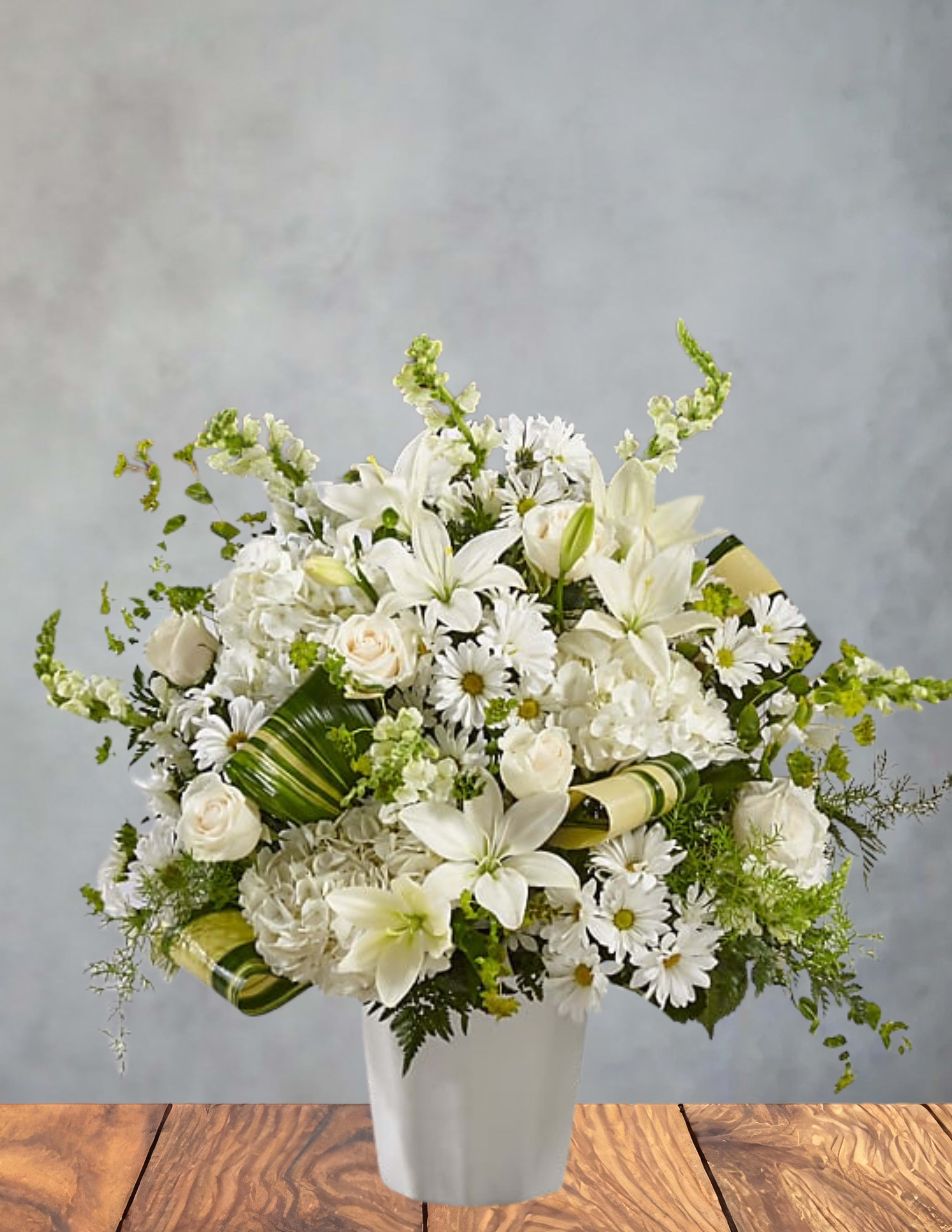  White Serenity - A luminous cluster of white lilies, roses, daisies and hydrangeas creates an immediate sense of calm-perfect for a sympathy arrangement, wedding centerpiece, or any moment when you want someone to feel deeply comforted. White Serenity is a formal white flower arrangement designed to read as both fresh and refined: the rounded composition gives a full, elegant silhouette while a few stems extend outward to keep the design feeling airy and natural.  This bouquet sits in a simple white vase tied with a soft green decorative ribbon, letting the white blooms take center stage while the pale green filler flowers and ribbon provide a gentle contrast. You'll find large hydrangea heads, fragrant lilies, classic white roses and cheerful daisy-like blooms arranged densely for impact yet balanced in height and spread-ideal for display on a table, mantle, or beside a memorial. The overall size and wide silhouette make it a striking focal piece without feeling overbearing.  For a grieving friend, a bride seeking understated elegance, or anyone who deserves a serene, beautiful reminder you're thinking of them, this arrangement reads as thoughtful and composed. Standard care-fresh water and a cool spot away from direct sun-will keep the flowers looking their best; ordering details and available delivery options are shown at checkout.