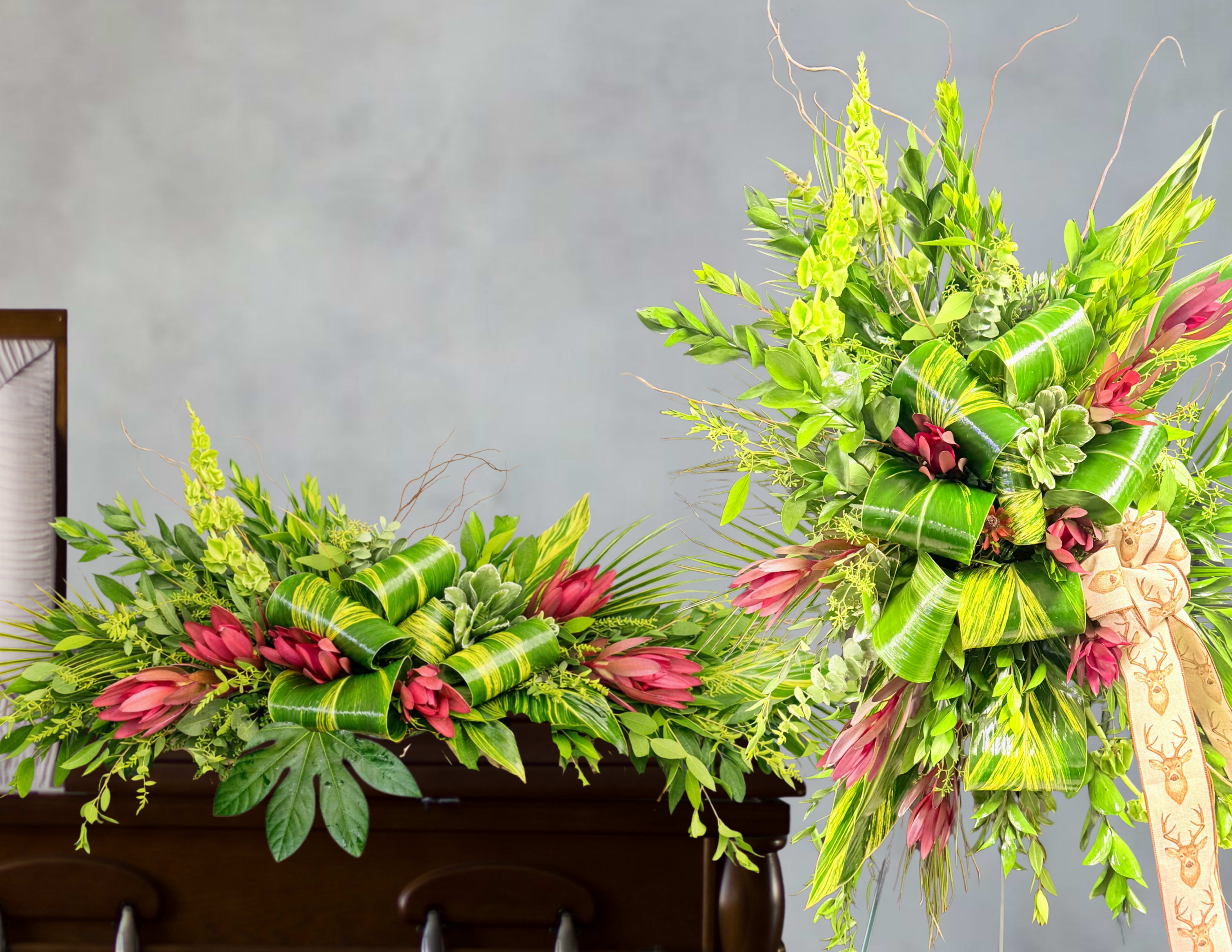 A life well rooted funeral service set  - Lush green foliage takes center stage in this funeral service set, paired with red accents to create a warm, celebratory tribute. This two-piece arrangement - one low and sprawling, the other taller and vertical on a stand - offers a balanced presentation that honors a life while conveying comfort and vibrancy. The tropical styling gives the display an uplifting presence that reads both respectful and life-affirming for a memorial service.  Layered broad glossy leaves, curled ribbon-like foliage, and airy branching elements build dramatic texture and depth, while pops of pink and touches of white bring tenderness and softness to the composition. Finished with tasteful ribbon and mounted on a supportive stand for the taller piece, the set is designed to be seen from multiple angles and to hold its shape throughout the service. Ideal for celebrating a loved one who cherished bold color and natural beauty, this arrangement arrives ready to display; contact us for delivery or sizing options to suit the service.