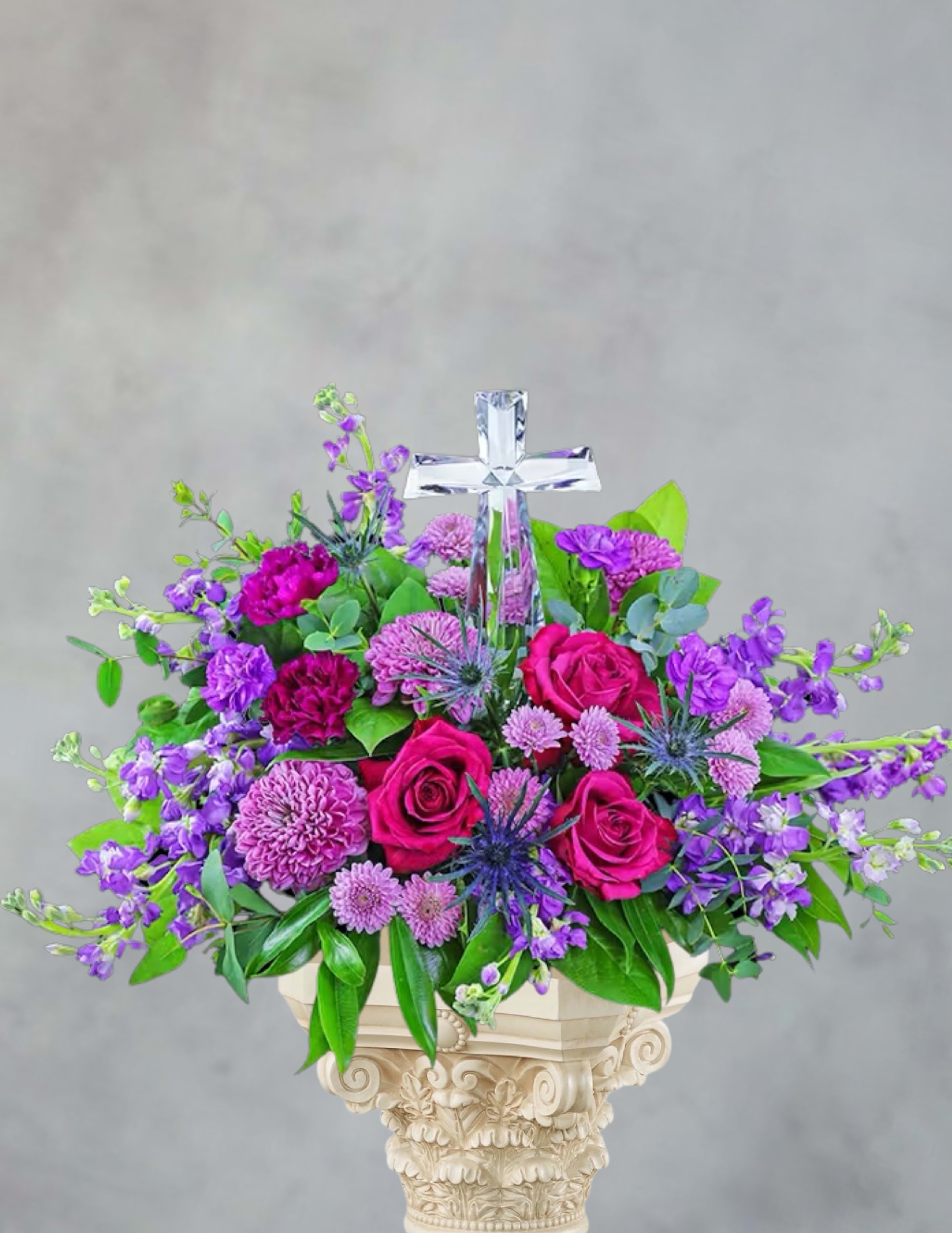 Purple and pink floral arrangement with a clear cross topper in a pedestal vase