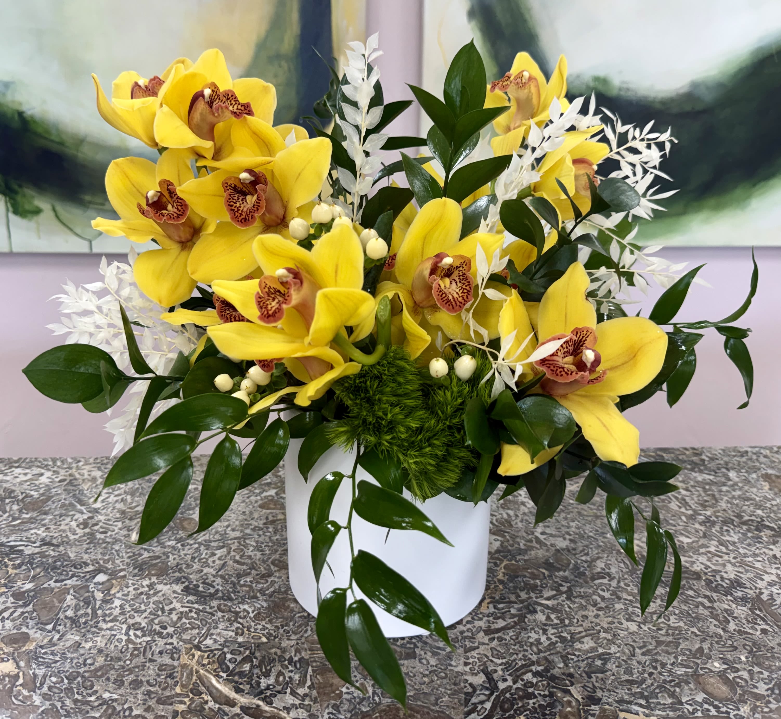 Orchid &amp; Vine Verdant - Clusters of bright yellow orchids with speckled centers make a cheerful statement in this flower arrangement-perfect as a birthday bouquet, a thank-you gift, or a bright pick-me-up for someone who appreciates tropical elegance. The orchid-forward design reads modern and intentional, so the person you're sending it to will feel genuinely celebrated and noticed the moment the box is opened.  The composition pairs those saturated yellow orchids with dark glossy foliage and delicate white accent sprays for contrast and texture; touches of soft pink and green keep the palette lively and layered. Arranged compactly in a clean white ceramic cylindrical vase, the piece reads polished and contemporary while the dense, rounded silhouette feels abundant without being fussy.  *Flowers are subject to substitutions of equal value.