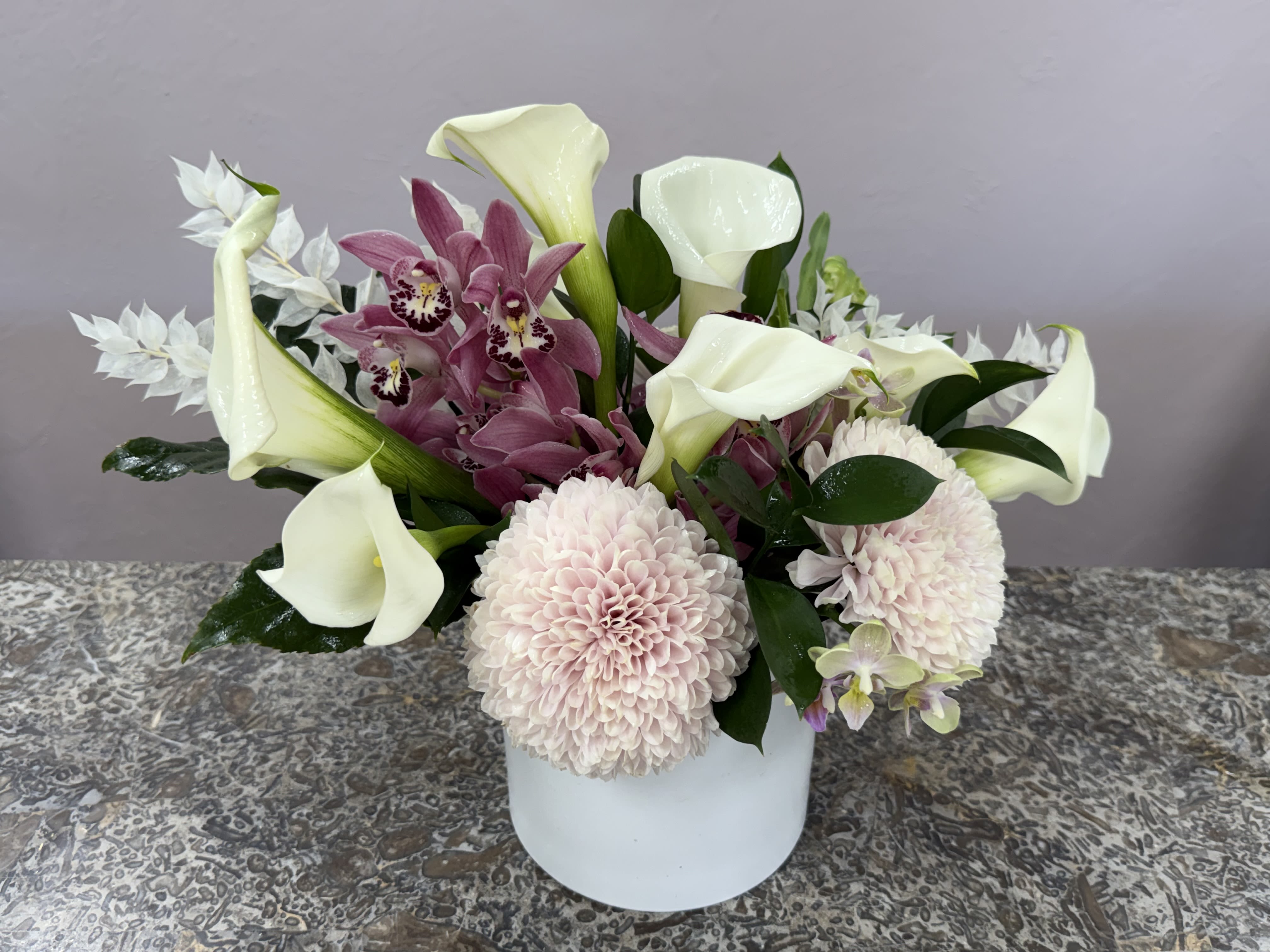 Calla and Orchid Study - A serene blend of white calla lilies and pink orchids creates an elegant arrangement perfect for a classy occasion or a refined everyday display. This low white ceramic vase holds a full composition that reads formal and gently romantic-she'll feel honored and comforted by the soft palette and sculptural lines of the lilies.  The bouquet mixes pure white calla lilies with clusters of pale pink orchids and blush chrysanthemums, nestled against glossy dark foliage for striking contrast and depth. The low, compact shape makes it ideal for tabletops or altars; the ceramic vessel keeps the look clean and modern while the textures-from the smooth calla spathes to the ruffled chrysanthemum petals-invite close appreciation.  Choose this arrangement when you want to convey sympathy, celebrate a wedding moment, or give someone a composed, sophisticated gift that makes them feel truly seen. The low vase arrives ready to display; simple fresh-water care will help the blooms stay graceful through the occasion.  *Flowers are subject to substitutions of equal value.