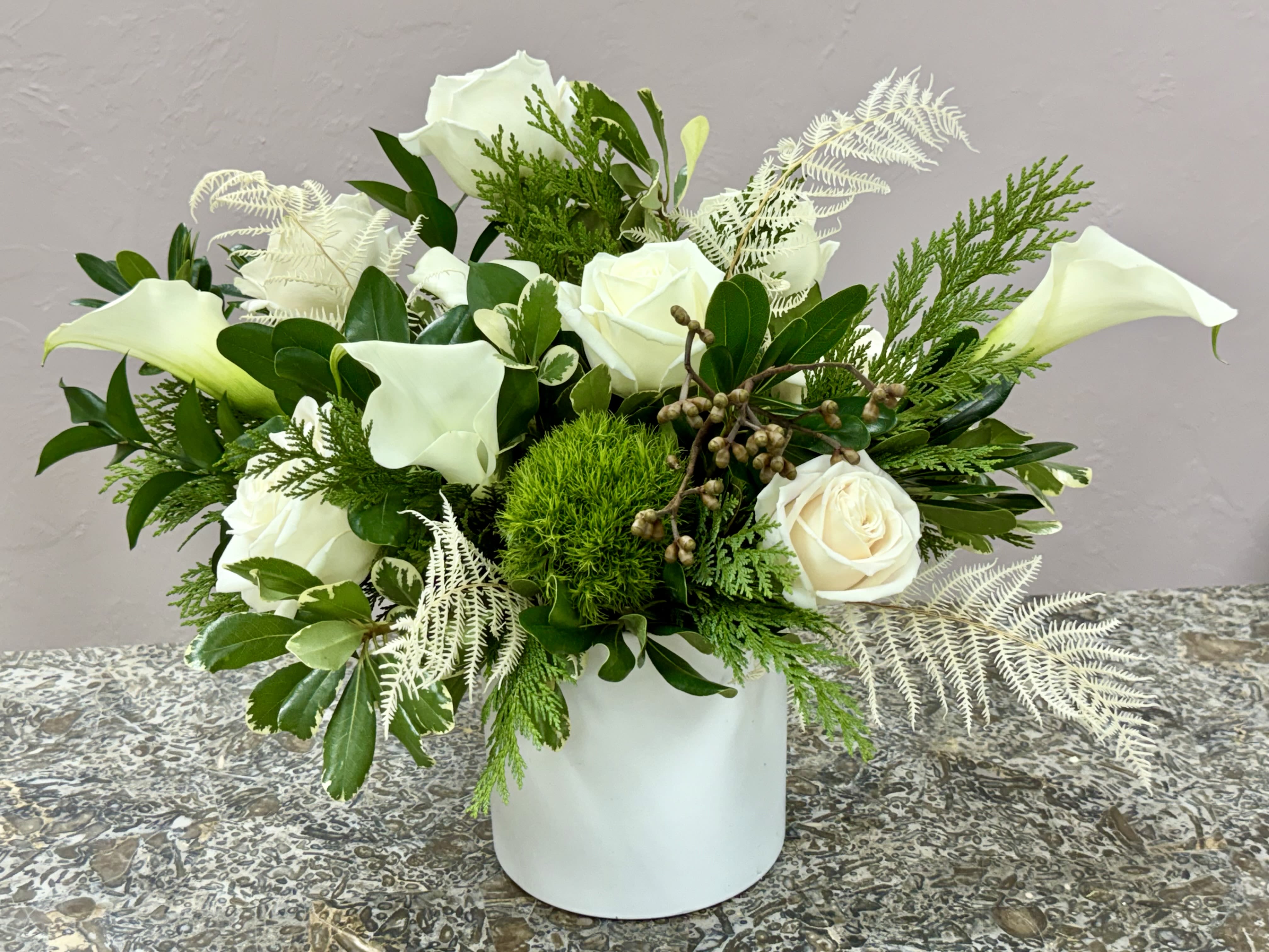 Naples Modern Ivory - Pure white calla lilies and creamy white roses form a calm, refined centerpiece perfect for a wedding table, sympathy arrangement, or as elegant everyday decor. The low, broadly spread silhouette gives the recipient a sense of thoughtful restraint-this is the kind of bouquet that makes someone feel comforted and seen.  Arranged in a compact white ceramic vase, the flowers sit amid dense mixed greenery and light, fern-like white accents that add soft texture and depth. The design reads medium-sized and low to moderately upright, with a layered, textured look that showcases the smooth calla stems and the lush rose heads in a mostly white-and-green palette.  Ideal for formal occasions or to bring quiet beauty into a home or office, this arrangement arrives ready to display-no extra trimming required beyond fresh water every few days. Its understated elegance makes it a meaningful way to mark weddings, sympathy, or to say you're thinking of someone in a tasteful, lasting way.  *Flowers are subject to substitutions of equal value.