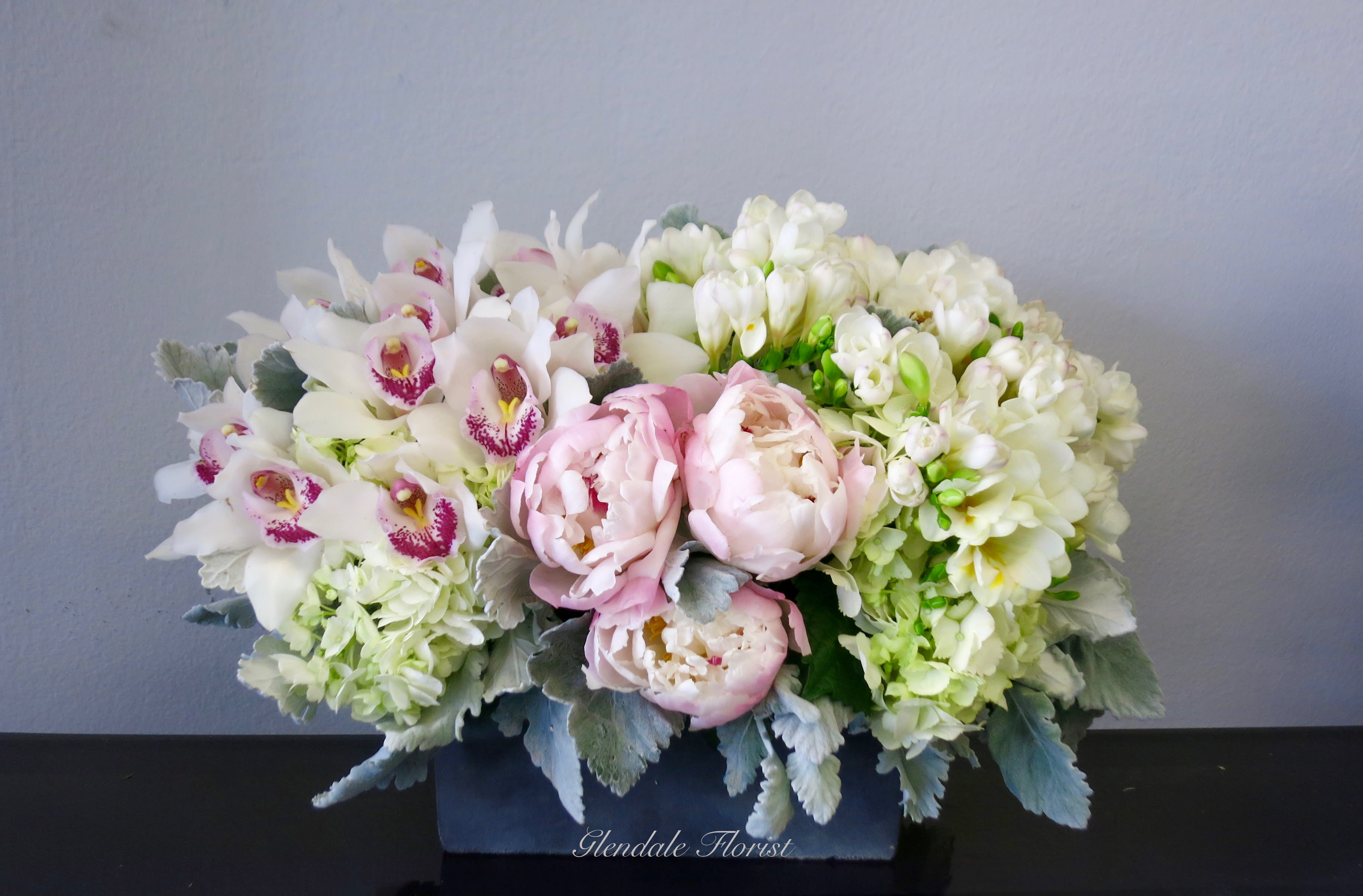 Low rectangular arrangement of white orchids, pale pink peonies, and cream flowers in a dark container