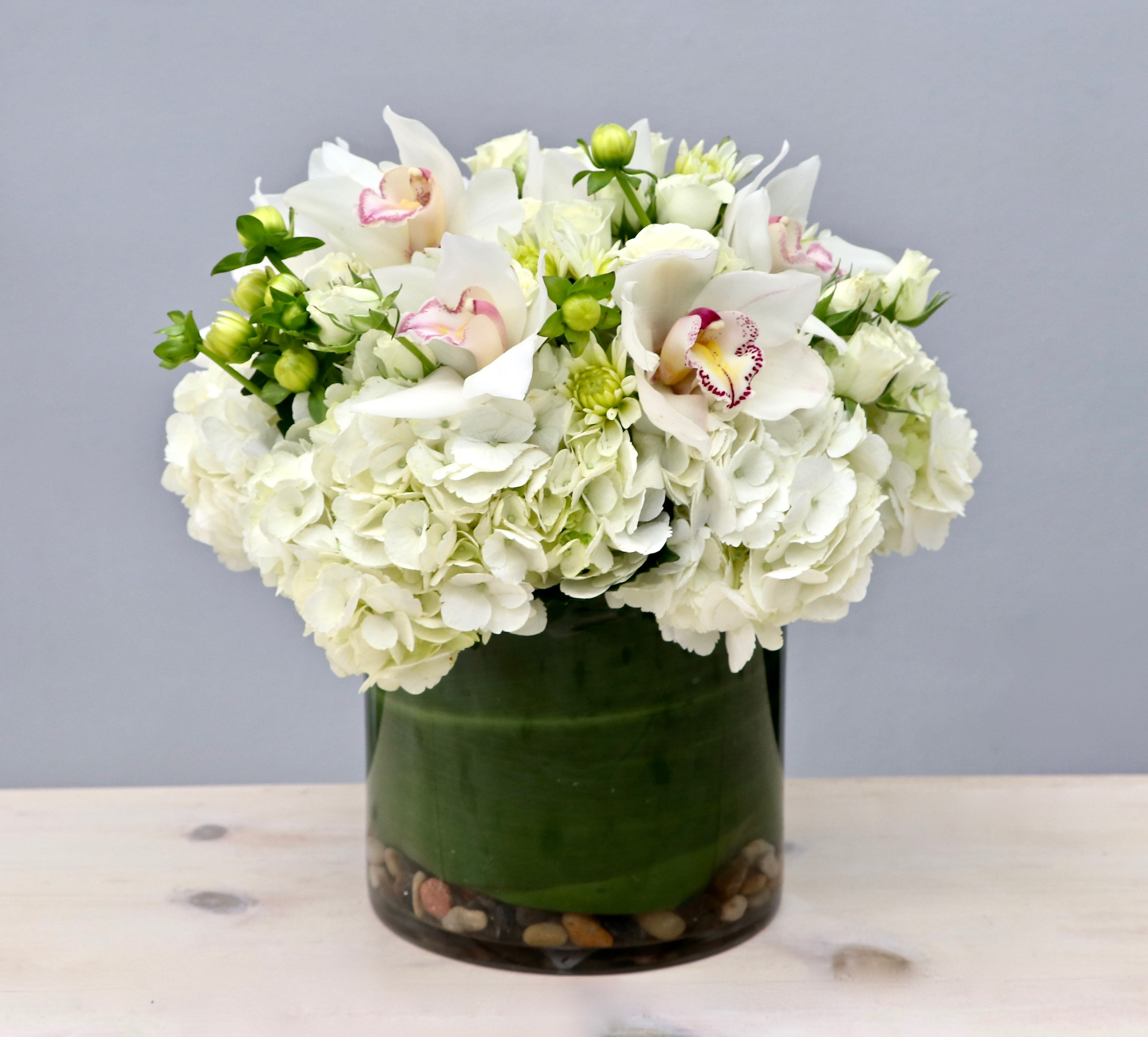 Low round arrangement of white hydrangeas and orchids in a glass cylinder vase with stones.