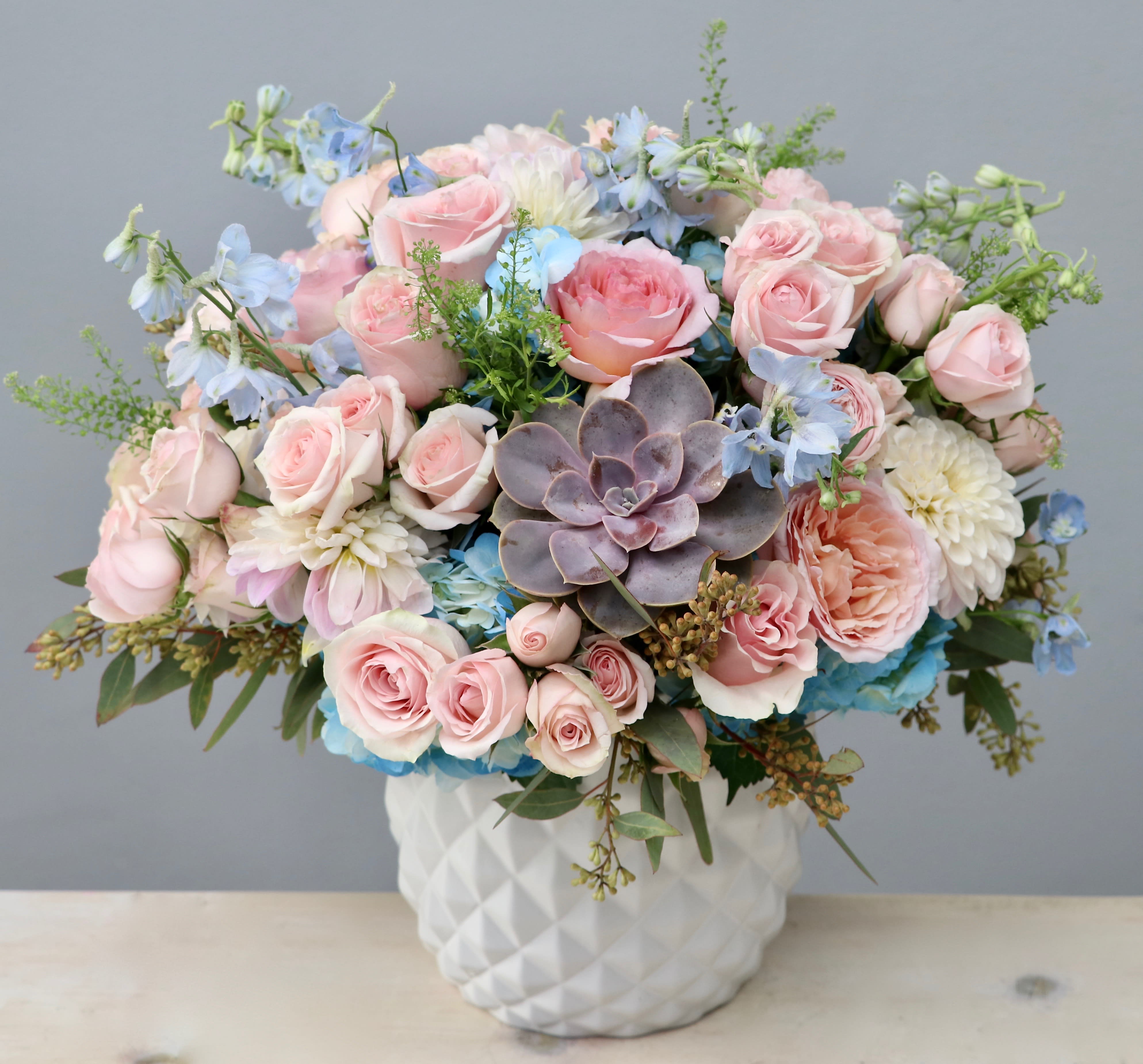 Pastel arrangement of pink roses, blue flowers, and a central succulent in a white ceramic vase