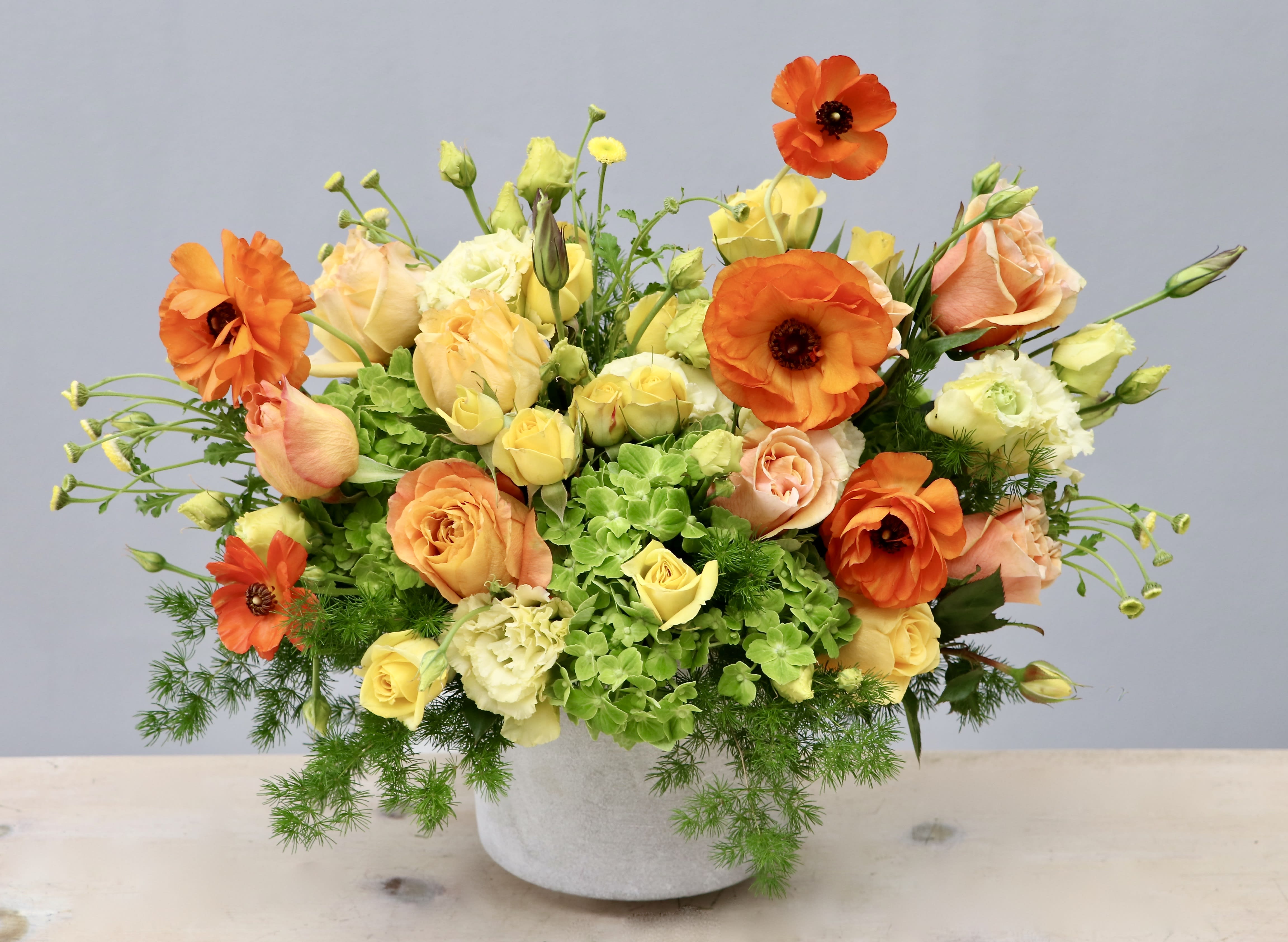 Low centerpiece of orange ranunculus, peach and yellow roses, and green hydrangeas in a white pot.