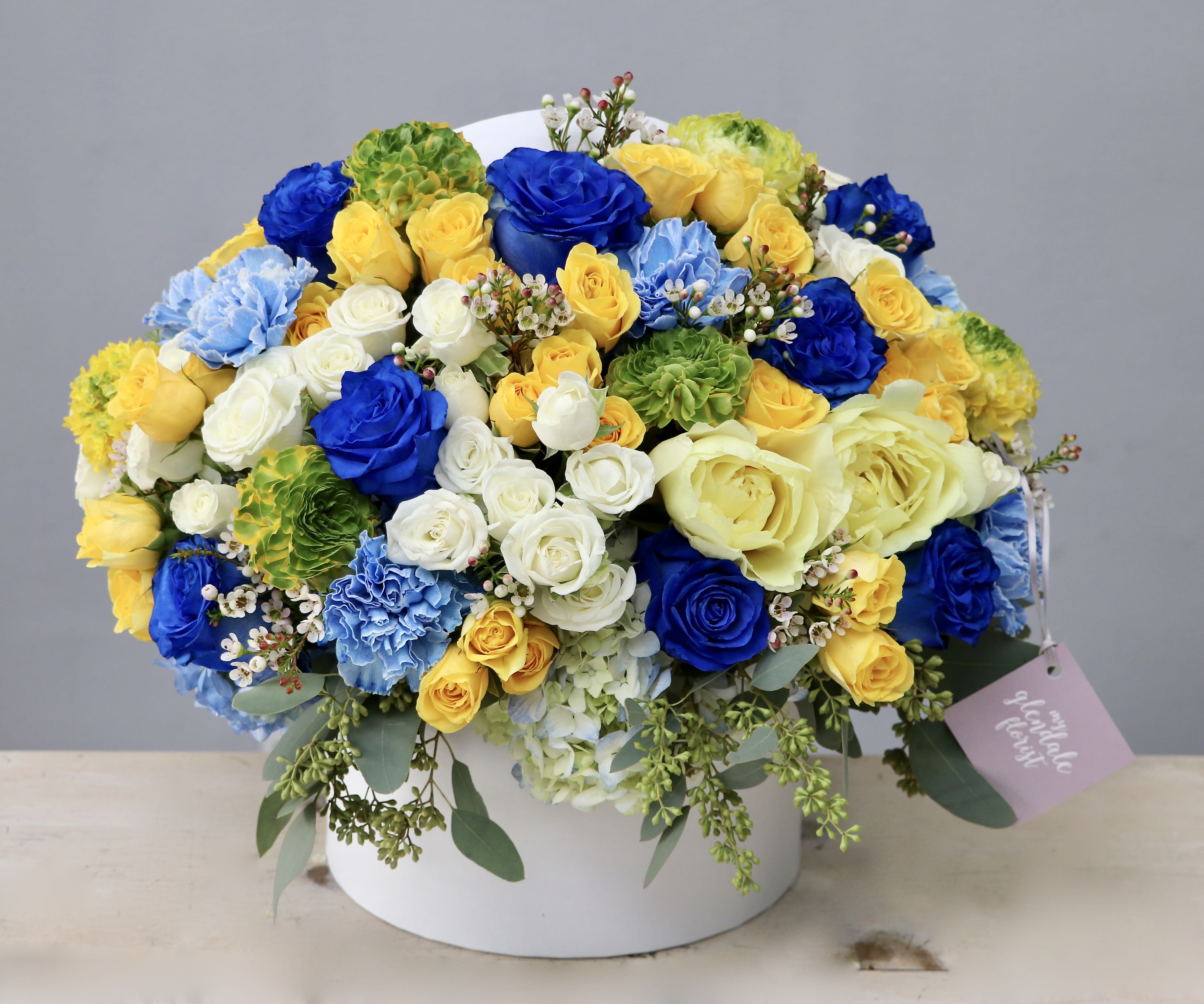 Round hatbox arrangement of yellow, blue, and white roses, carnations, and hydrangeas with a gift tag
