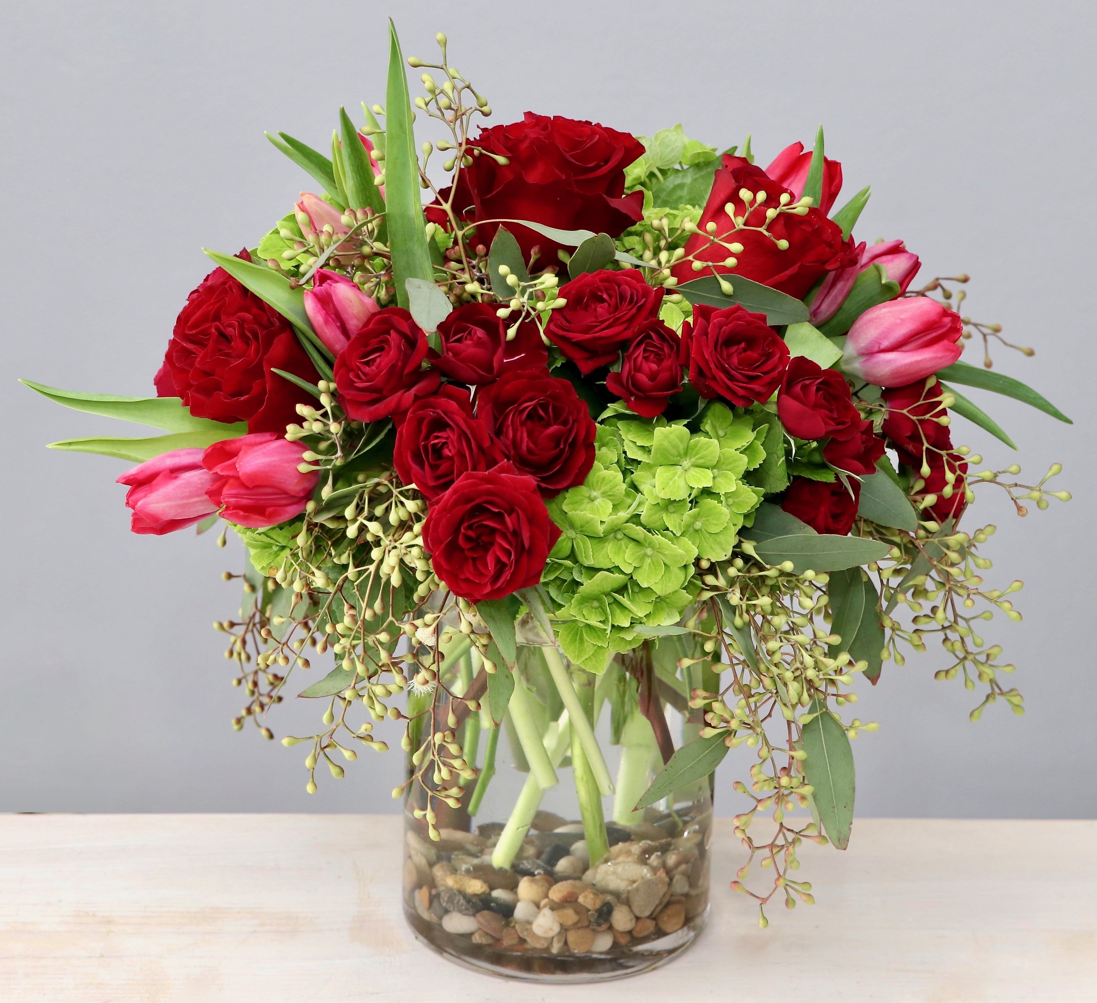 Arrangement of red roses, pink tulips, and green hydrangeas in a clear glass vase with river rocks