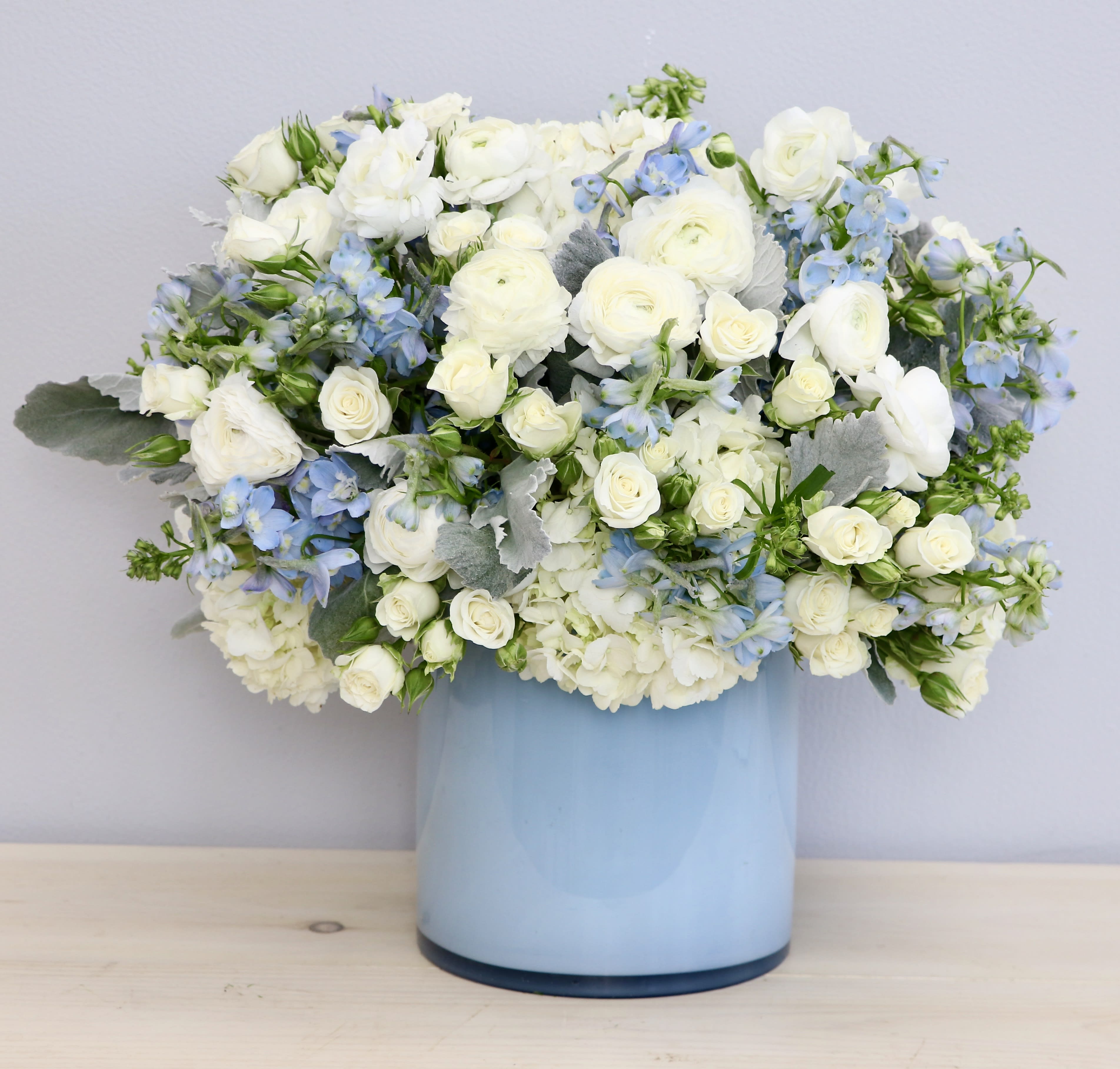 Round arrangement of white and light blue flowers in a light blue cylinder vase