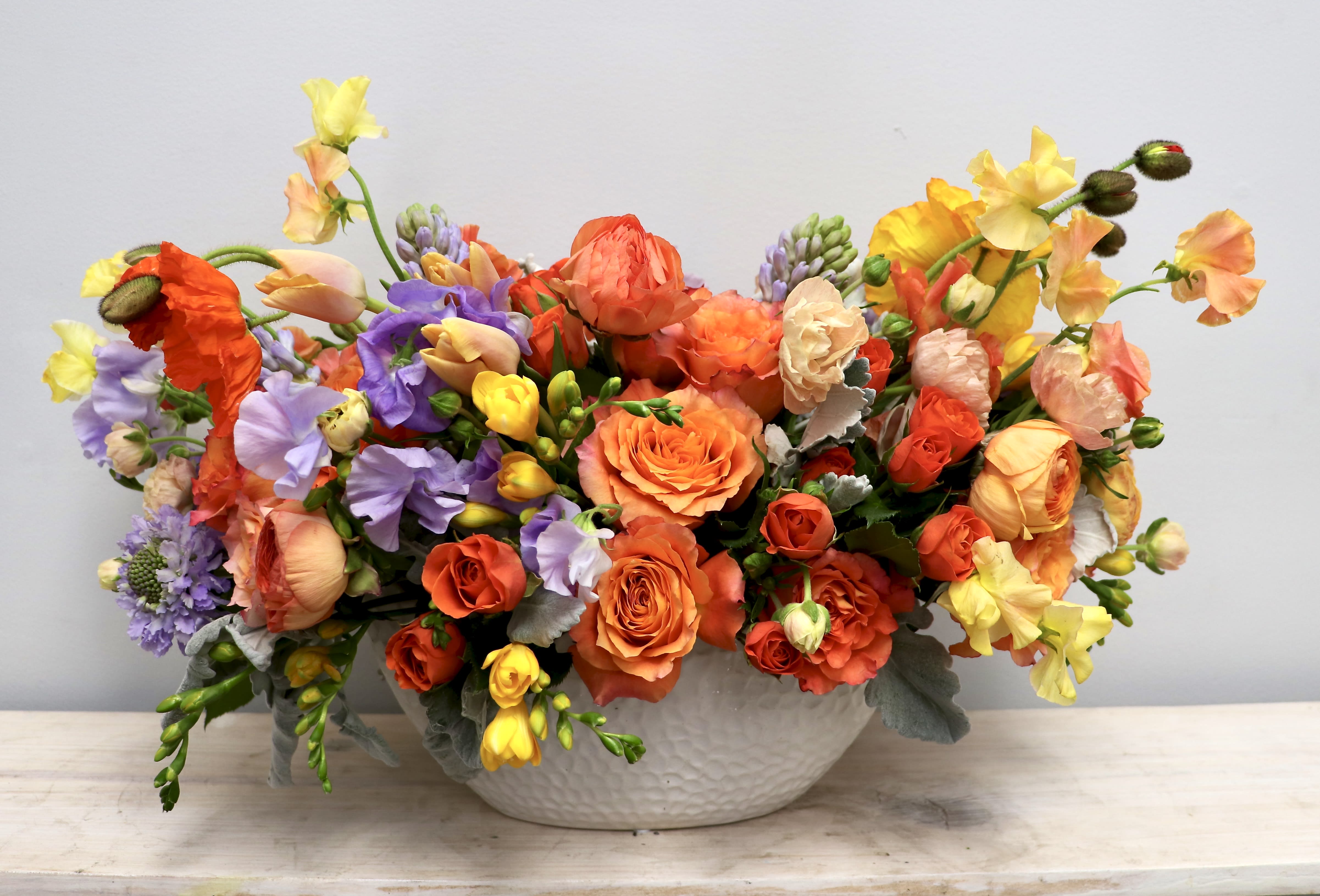 Low ceramic bowl filled with orange roses, ranunculus, and mixed yellow and lavender blooms in a lush arrangement