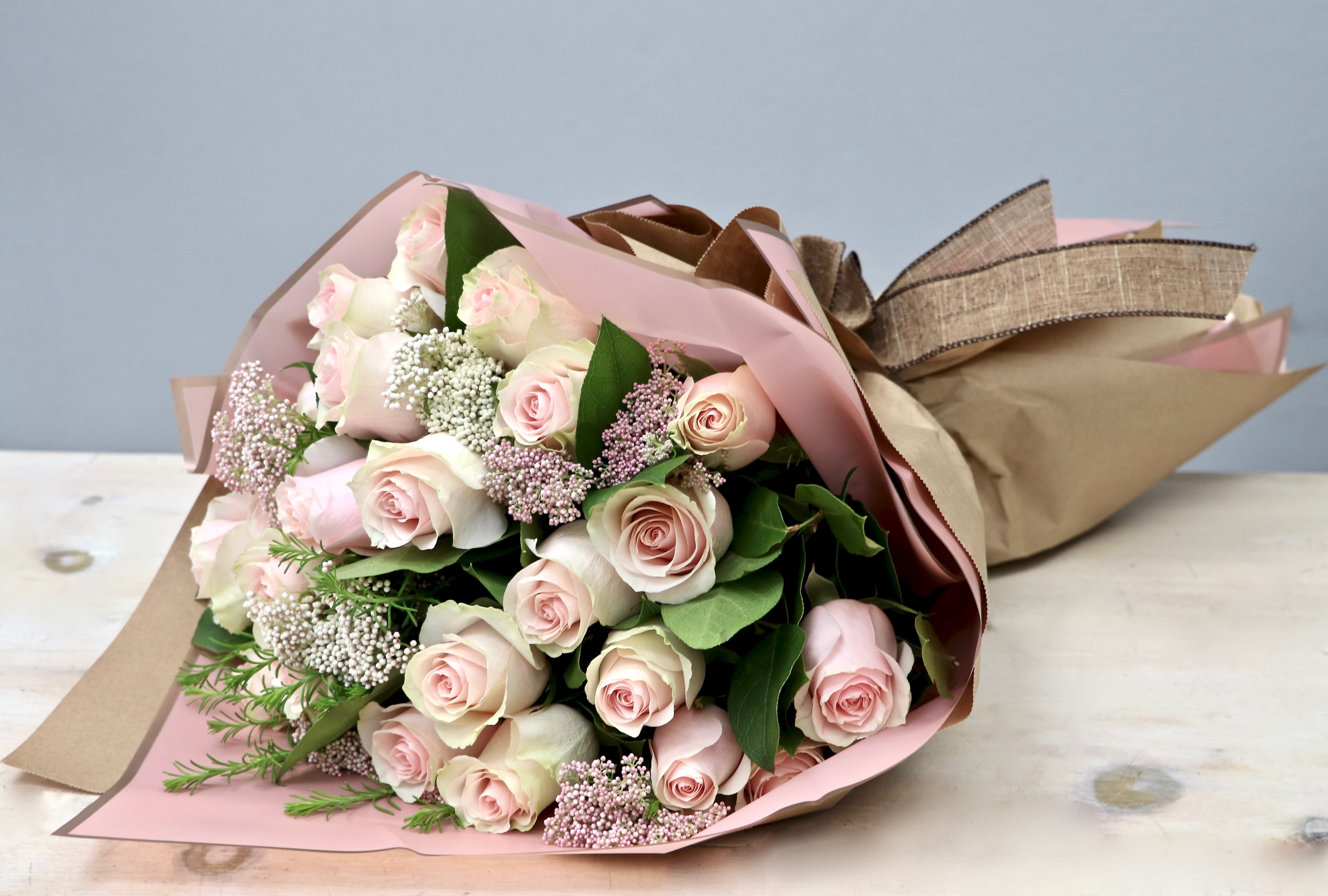 Hand-tied bouquet of pale pink roses with white filler flowers wrapped in pink and kraft paper