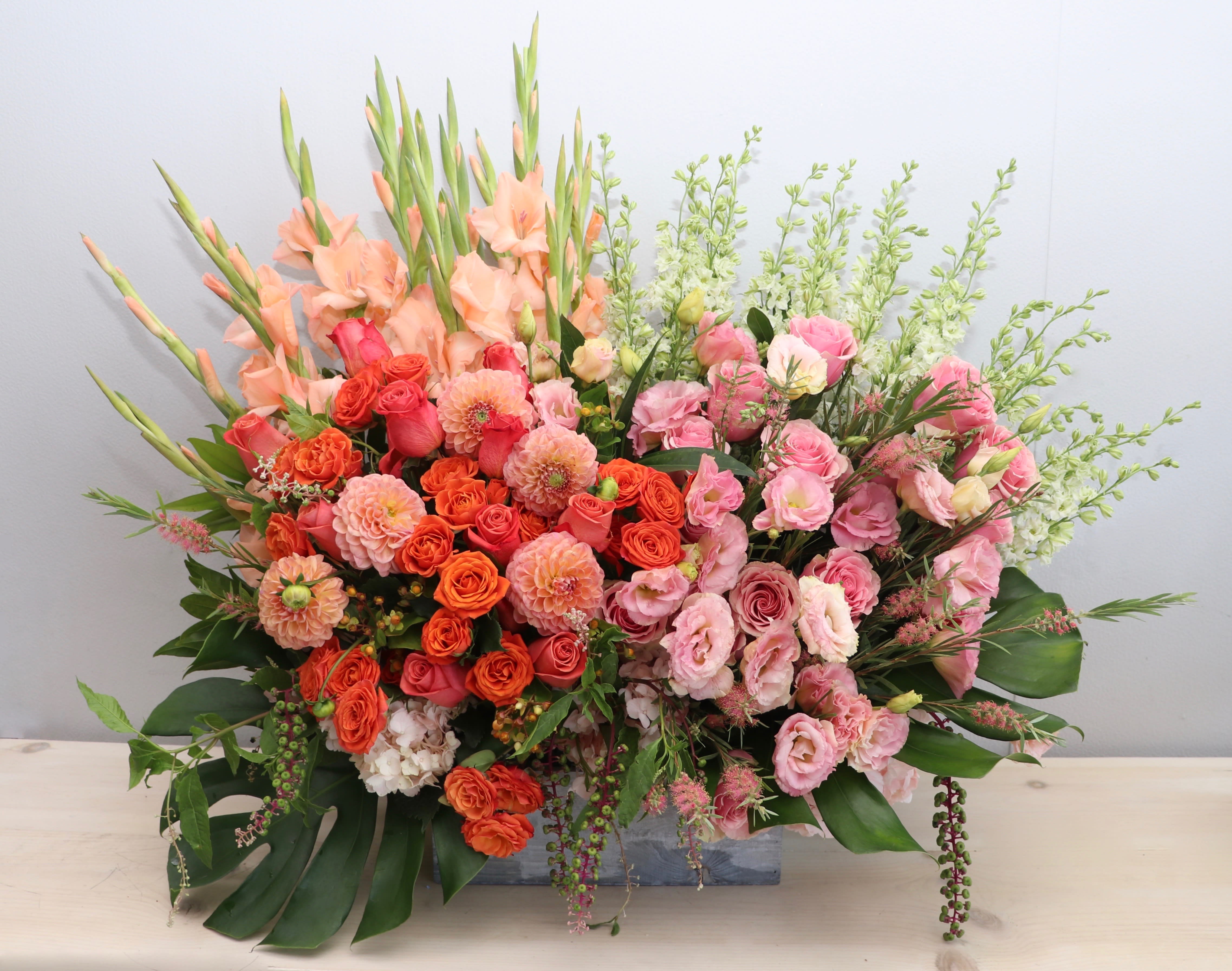 Large rectangular arrangement of peach gladiolus, orange dahlias, and pink roses in a low wooden container