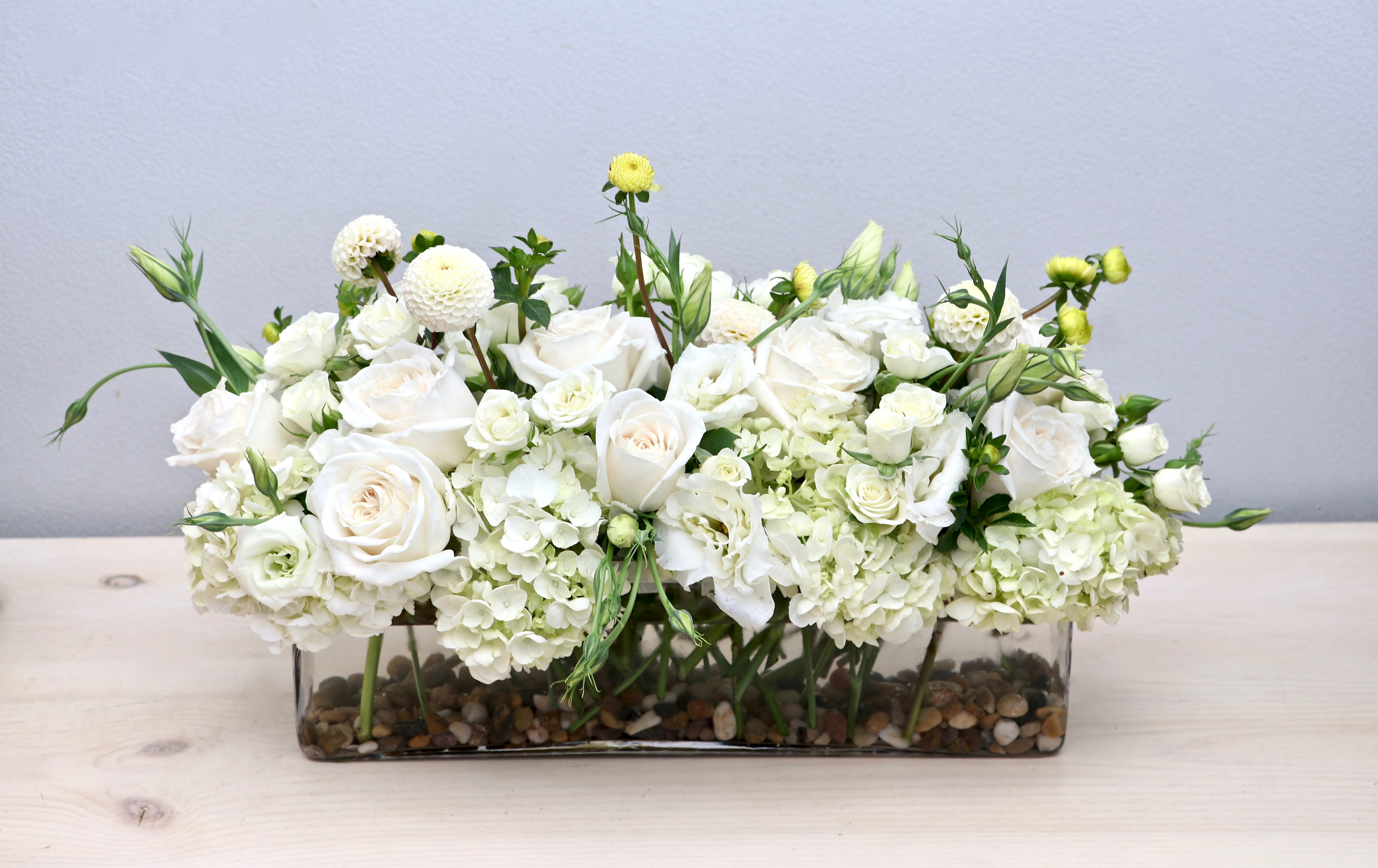 Low white rose and hydrangea arrangement in a clear rectangular vase with pebbles at the base