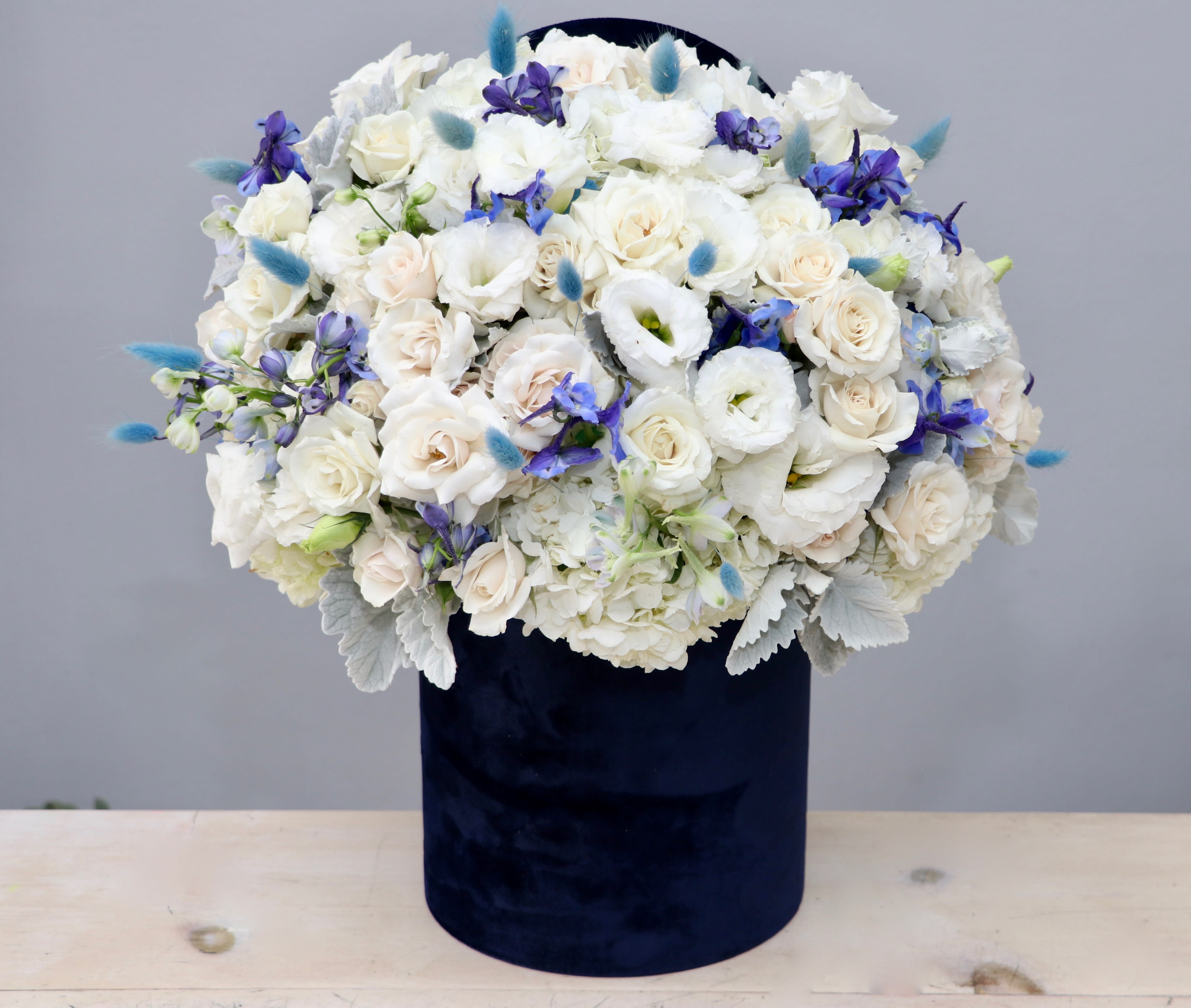 Round arrangement of white and cream flowers with blue accents in a tall navy velvet box