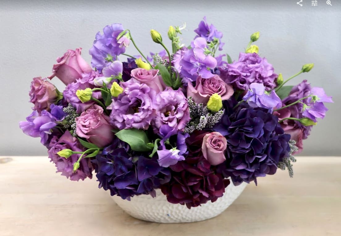 Low arrangement of purple and lavender roses and lisianthus in a white bowl vase
