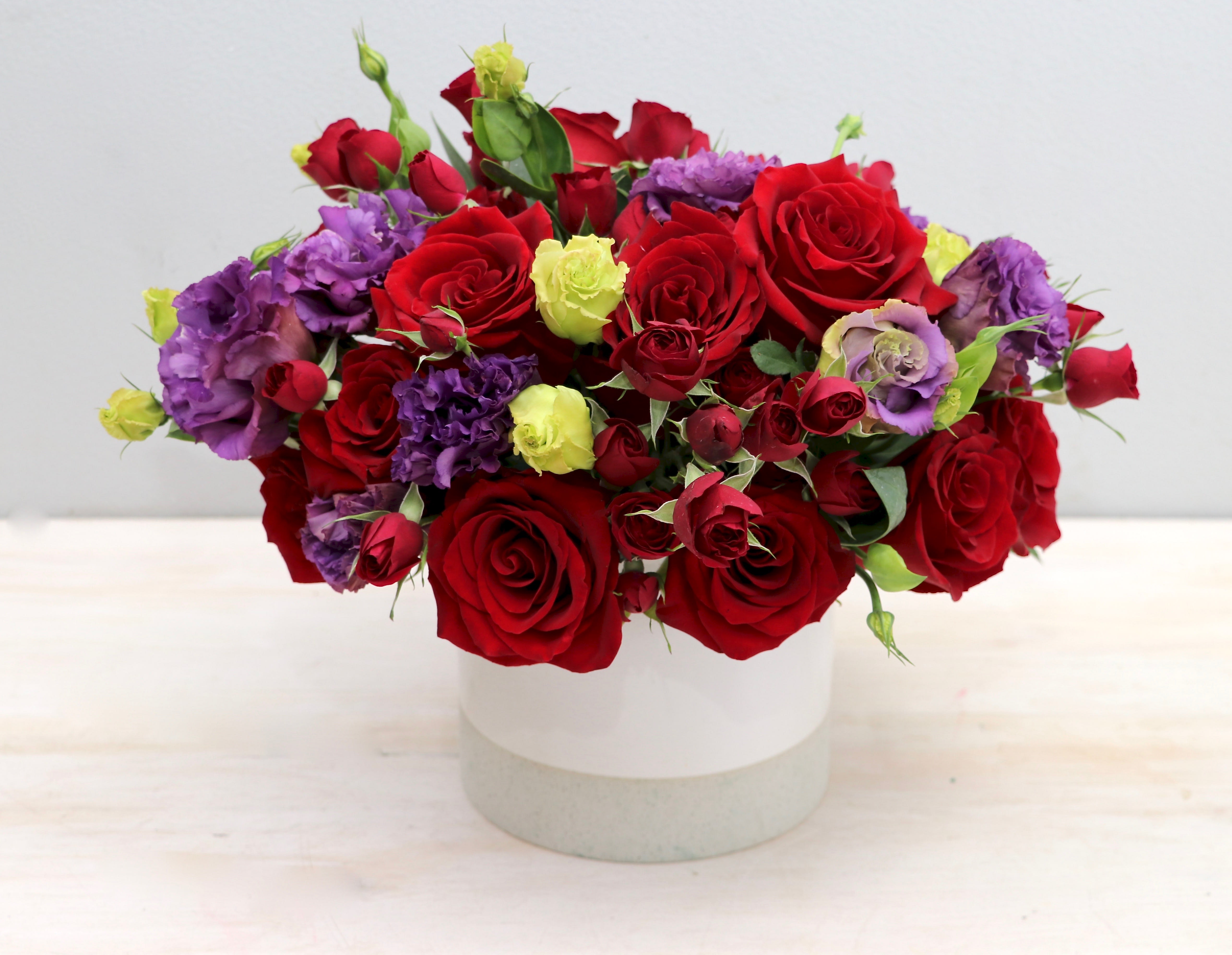 Low arrangement of red roses with purple and light green blooms in a white ceramic vase